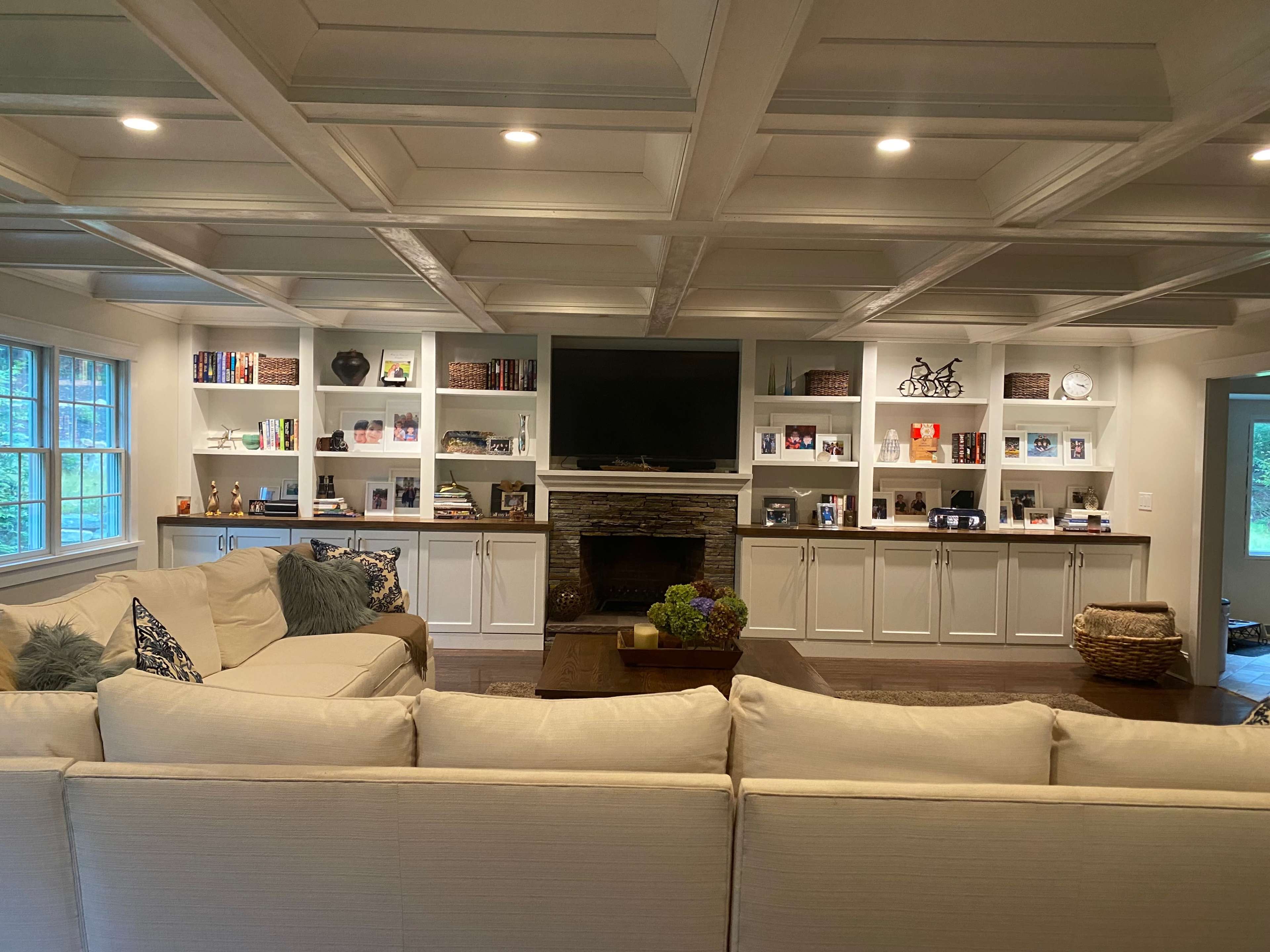 The room features a spacious living area with a sectional sofa facing a stone fireplace, built-in shelving filled with books and decorative items, and a wall-mounted television.