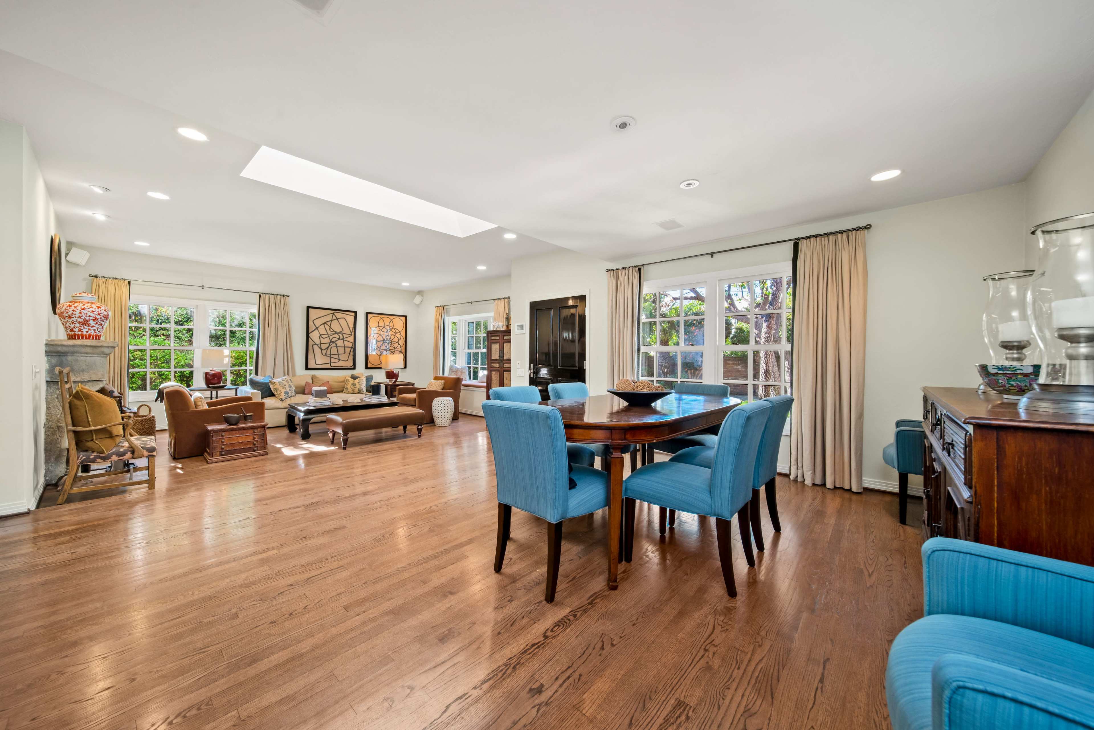 A spacious living room with hardwood floors, large windows, and a dining area featuring a round table surrounded by blue chairs.
