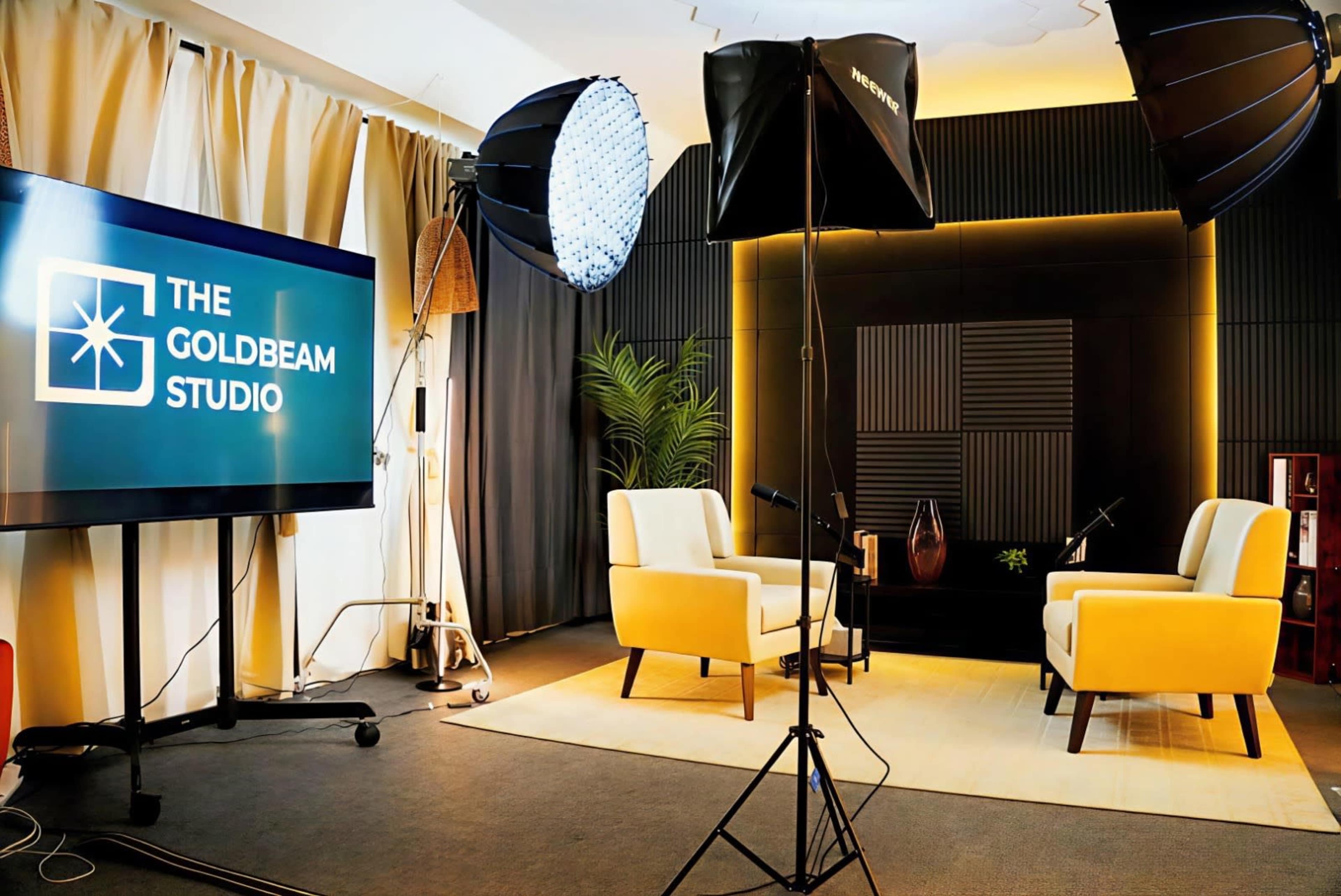 The image shows a well-lit studio setup with two yellow chairs facing a screen displaying "THE GOLDBEAM STUDIO," along with professional lighting equipment.