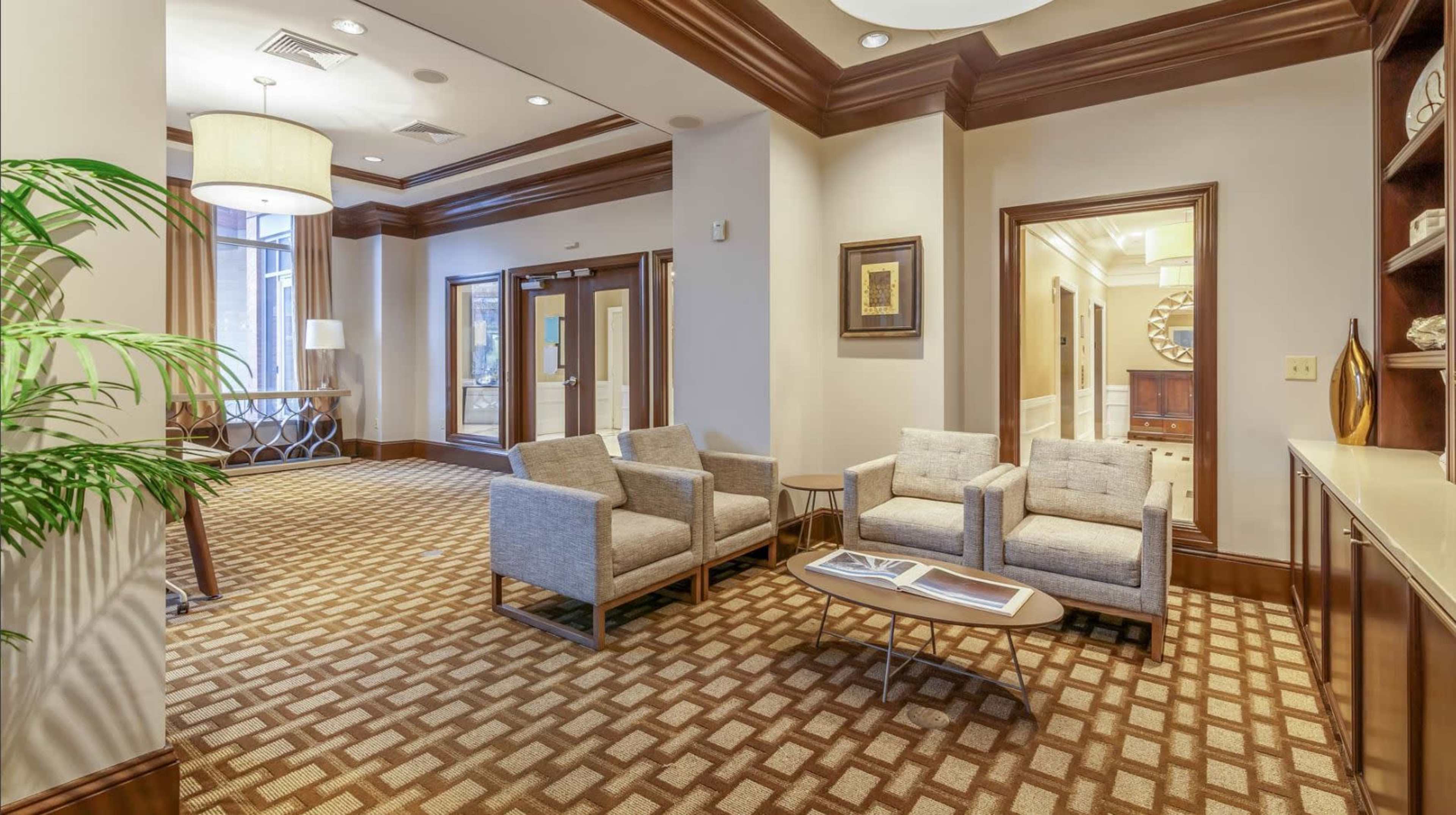 A modern lobby area featuring gray upholstered chairs, a wooden coffee table, and a decorative plant, with an entrance visible in the background.