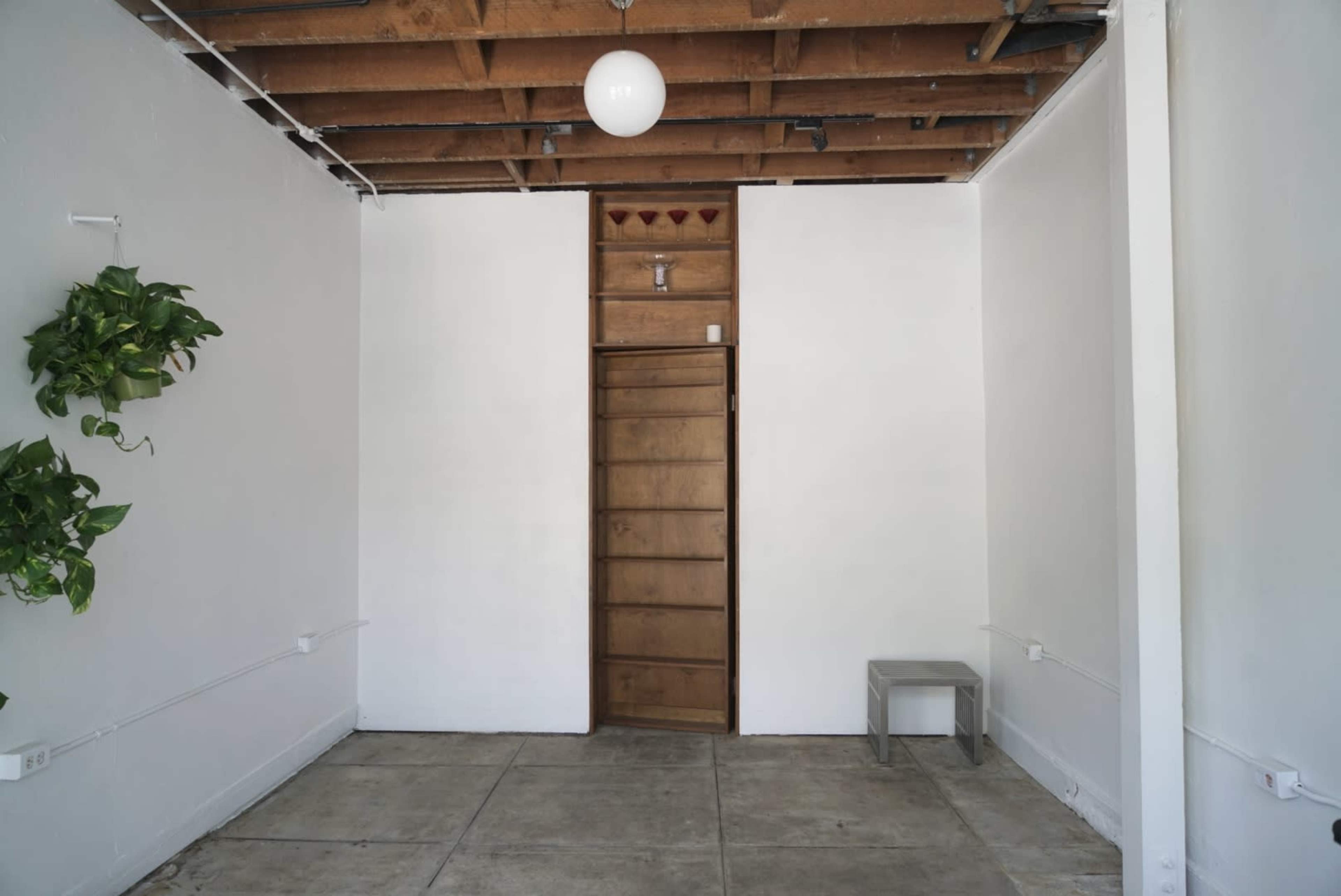 A sparsely furnished room with exposed wooden beams, a single overhead light, a door at the far end, and a potted plant on the left wall.