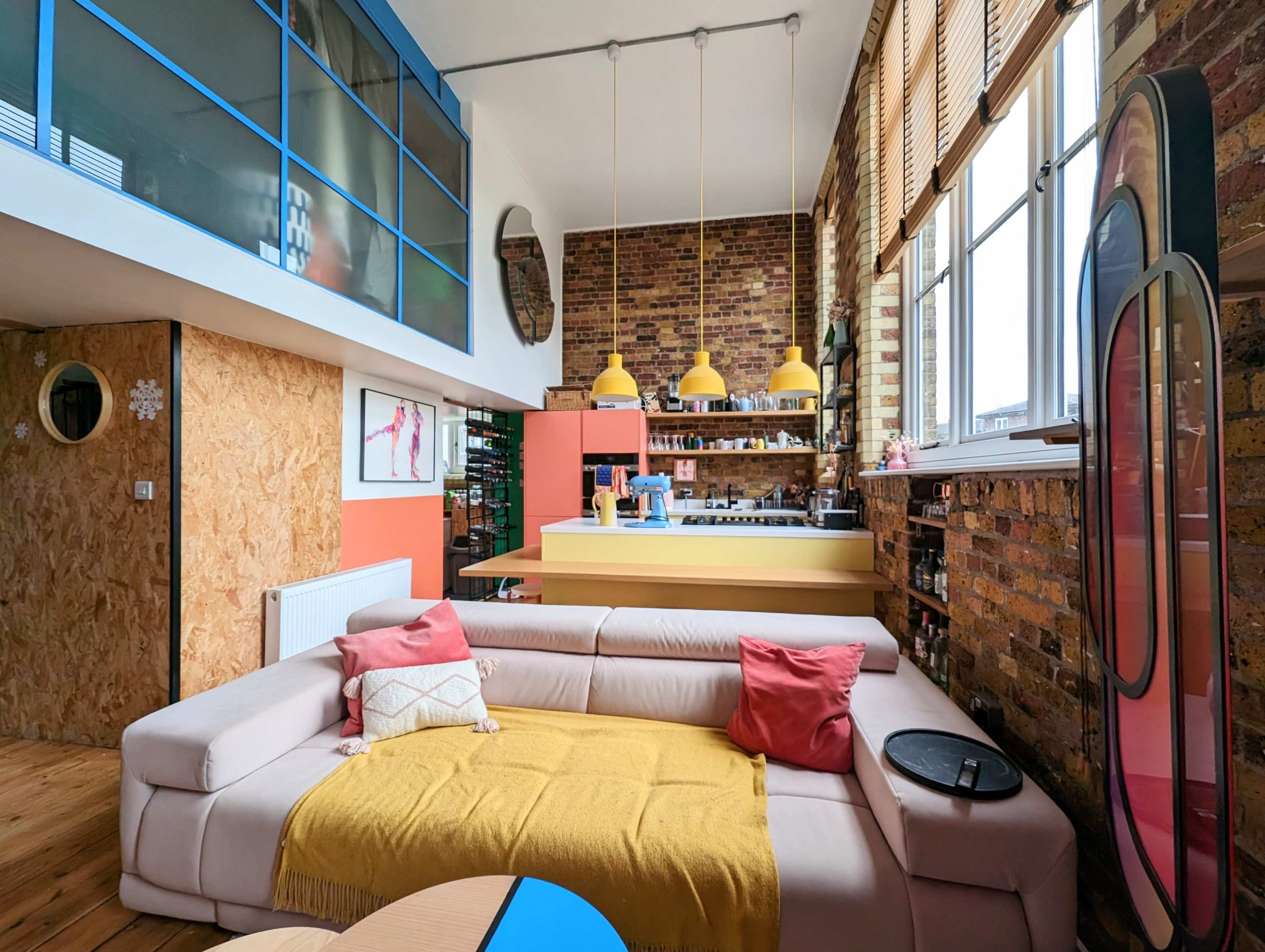 A modern living room features a large beige sofa with colorful cushions, a yellow throw, and a kitchenette with bright decor, all against a backdrop of exposed brick walls and large windows.