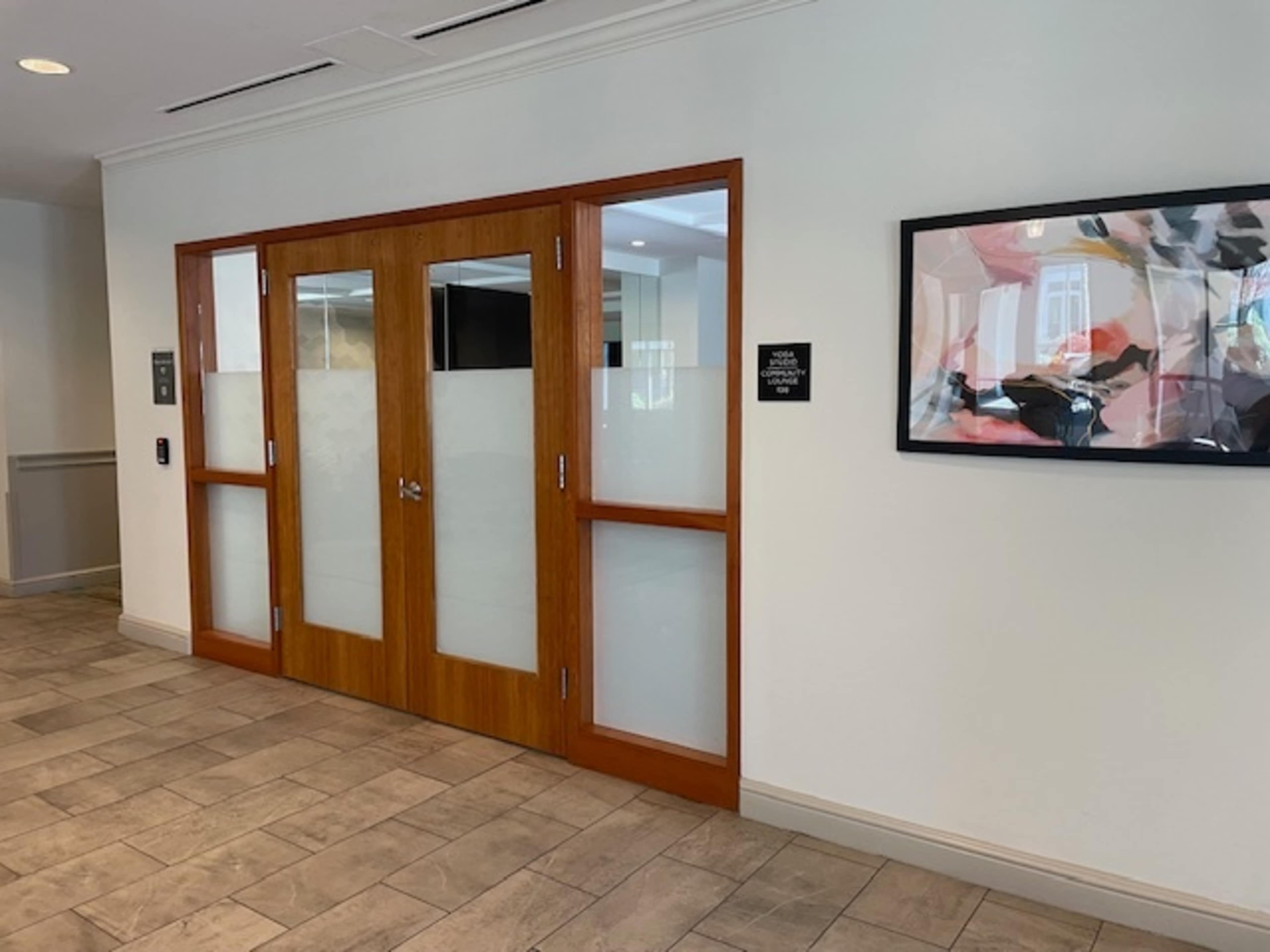 The image shows a wooden door with large glass panels, leading to a room, set within a modern interior hallway.