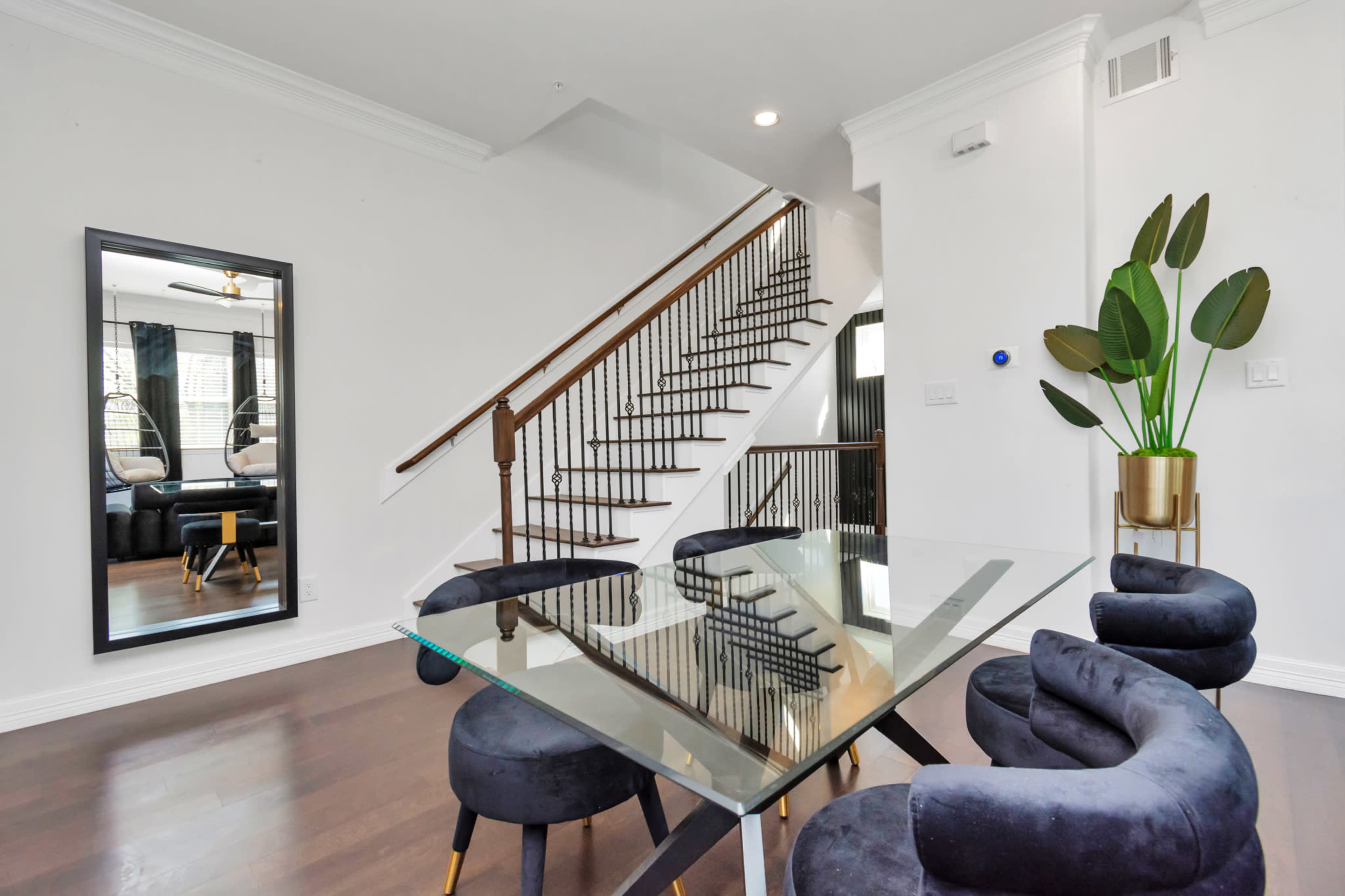 A modern dining area features a glass table surrounded by blue velvet chairs, with a staircase and a large mirror reflecting the space.