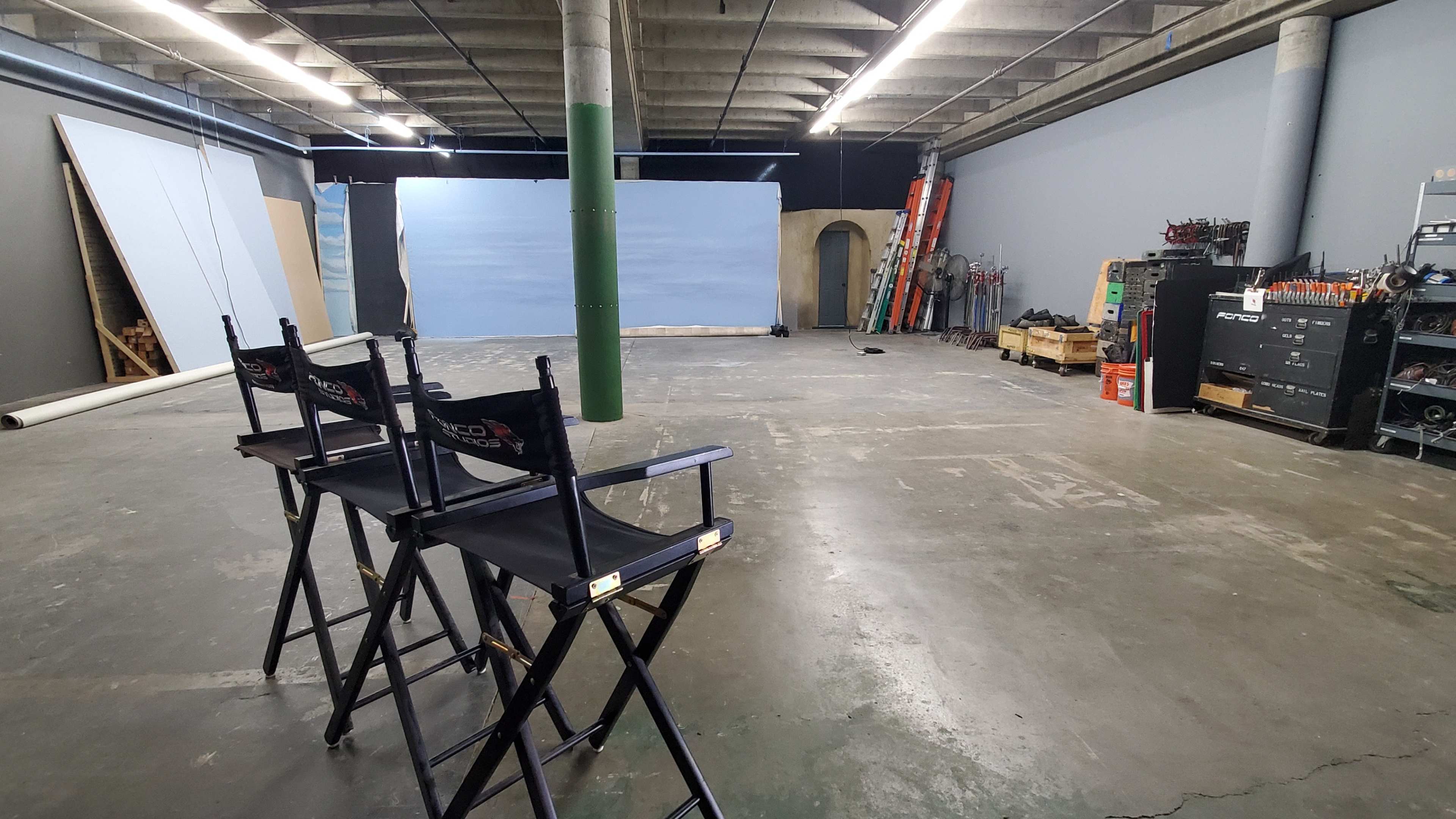 A vacant studio space featuring two director's chairs facing a backdrop, with equipment and props stored along the walls.