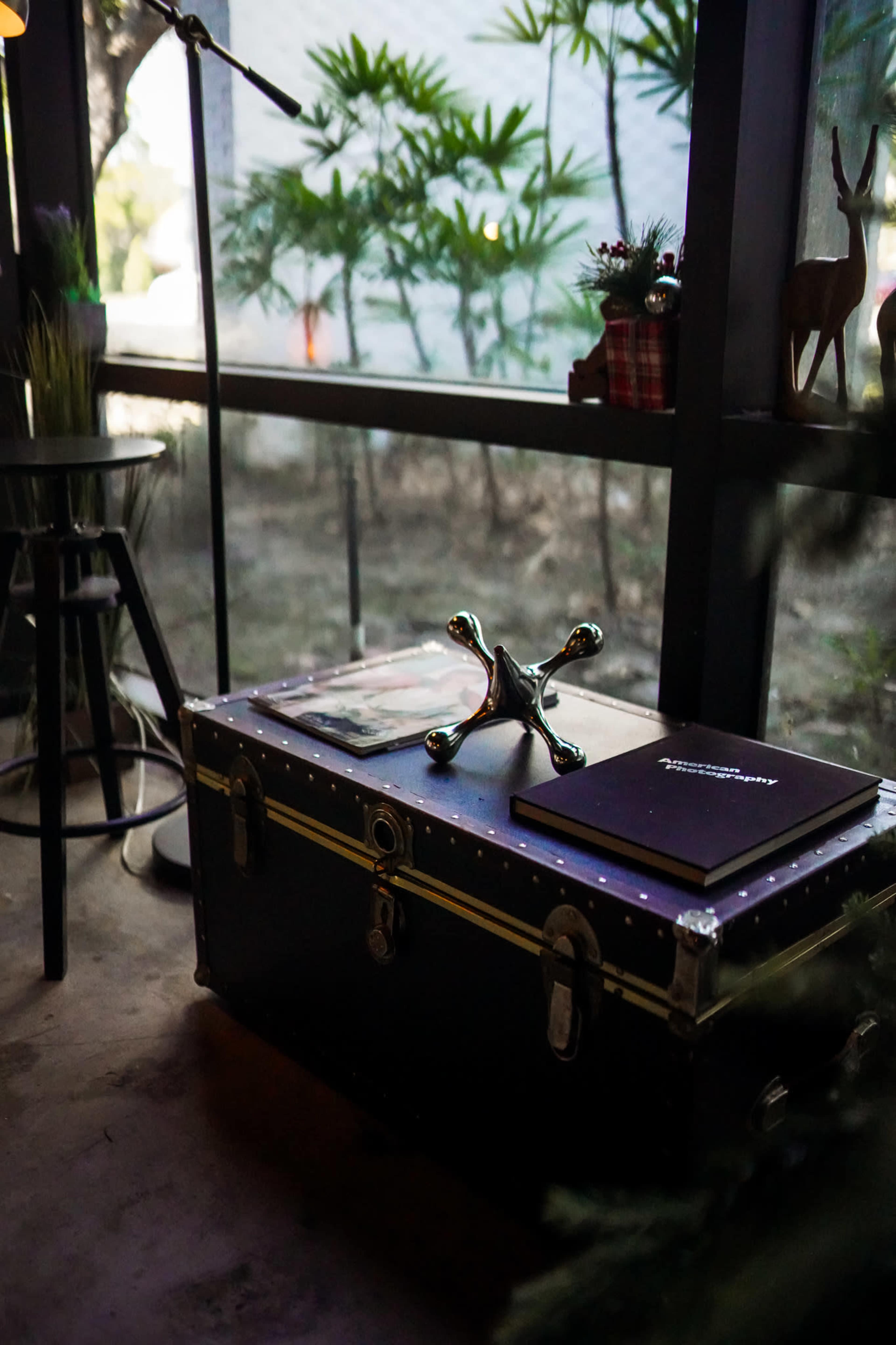 The image shows a vintage trunk placed beside a window, with a metal sculpture on top and plants visible in the background.