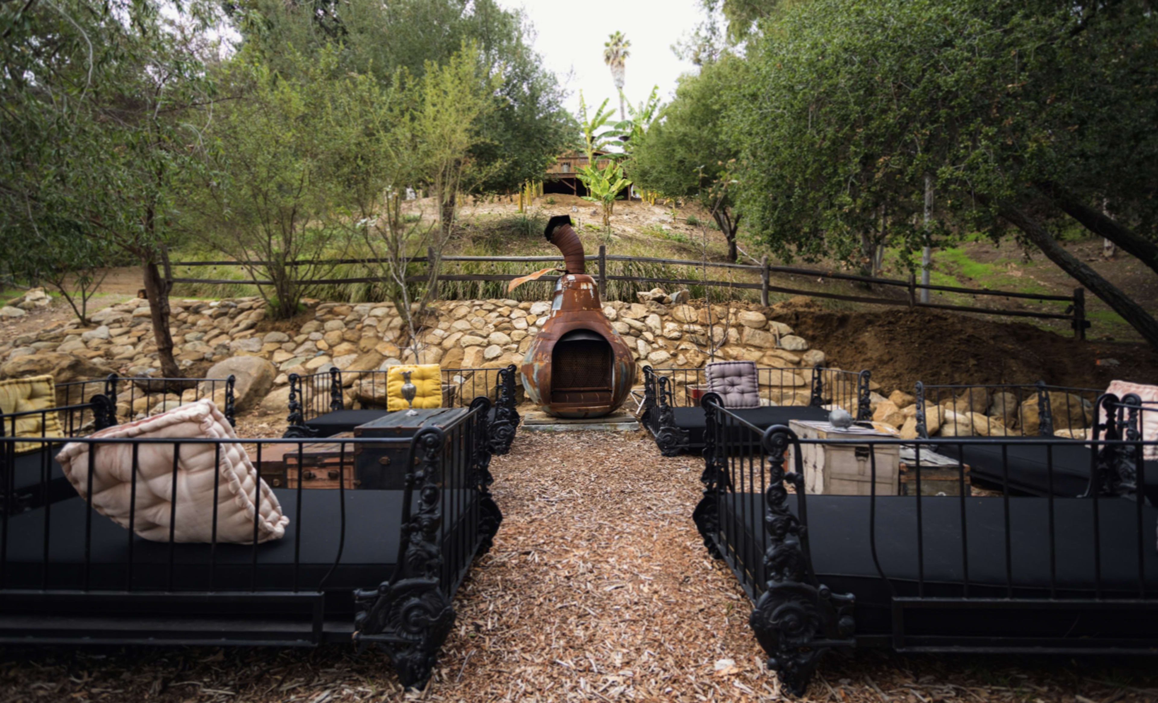 The scene features several black iron-frame beds arranged around a rustic outdoor fireplace, with a landscaped hillside and trees in the background.
