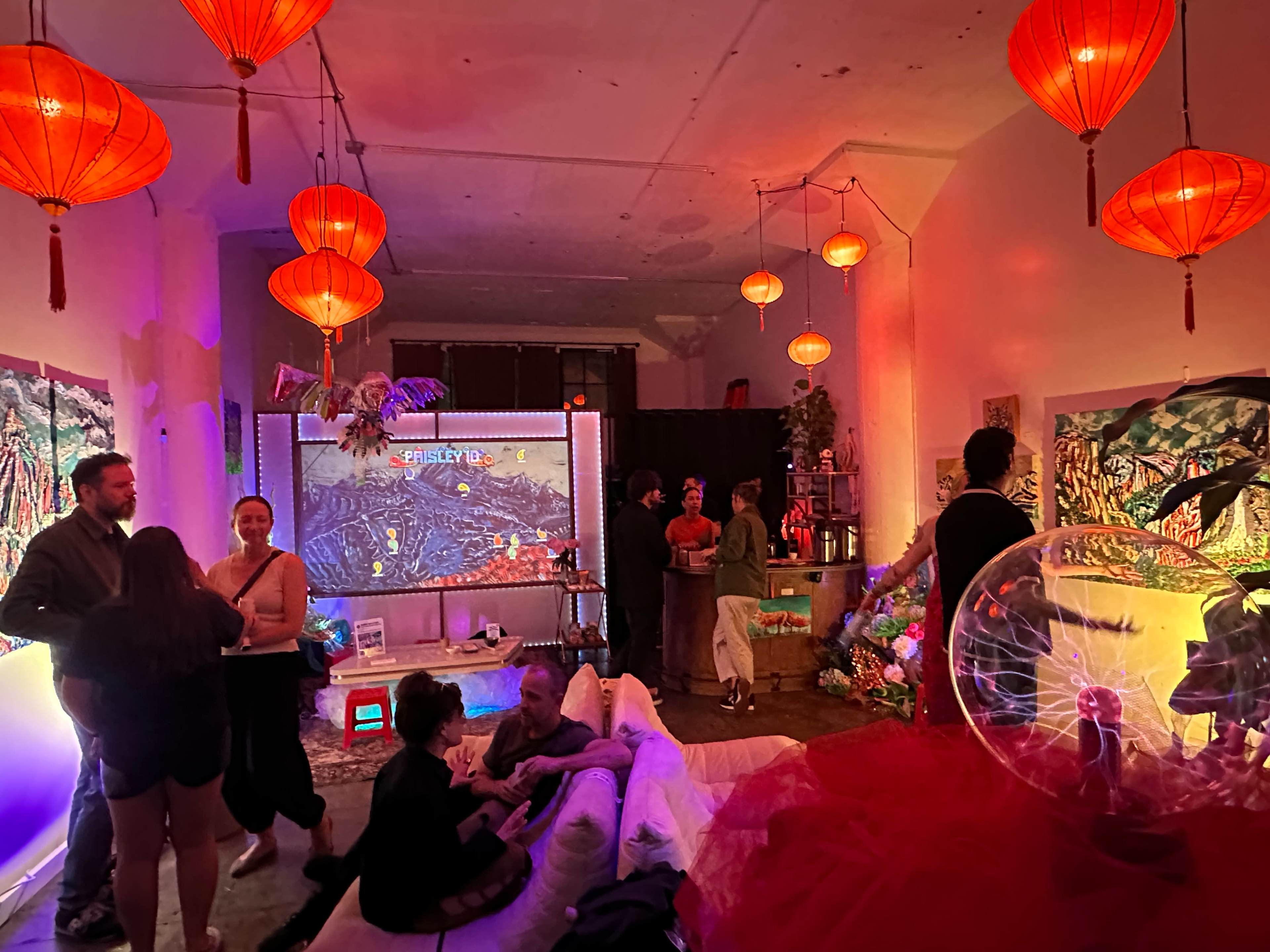 The image shows a vibrant, eclectic indoor space adorned with red lanterns, colorful artwork on the walls, and people socializing amidst an artistic atmosphere.
