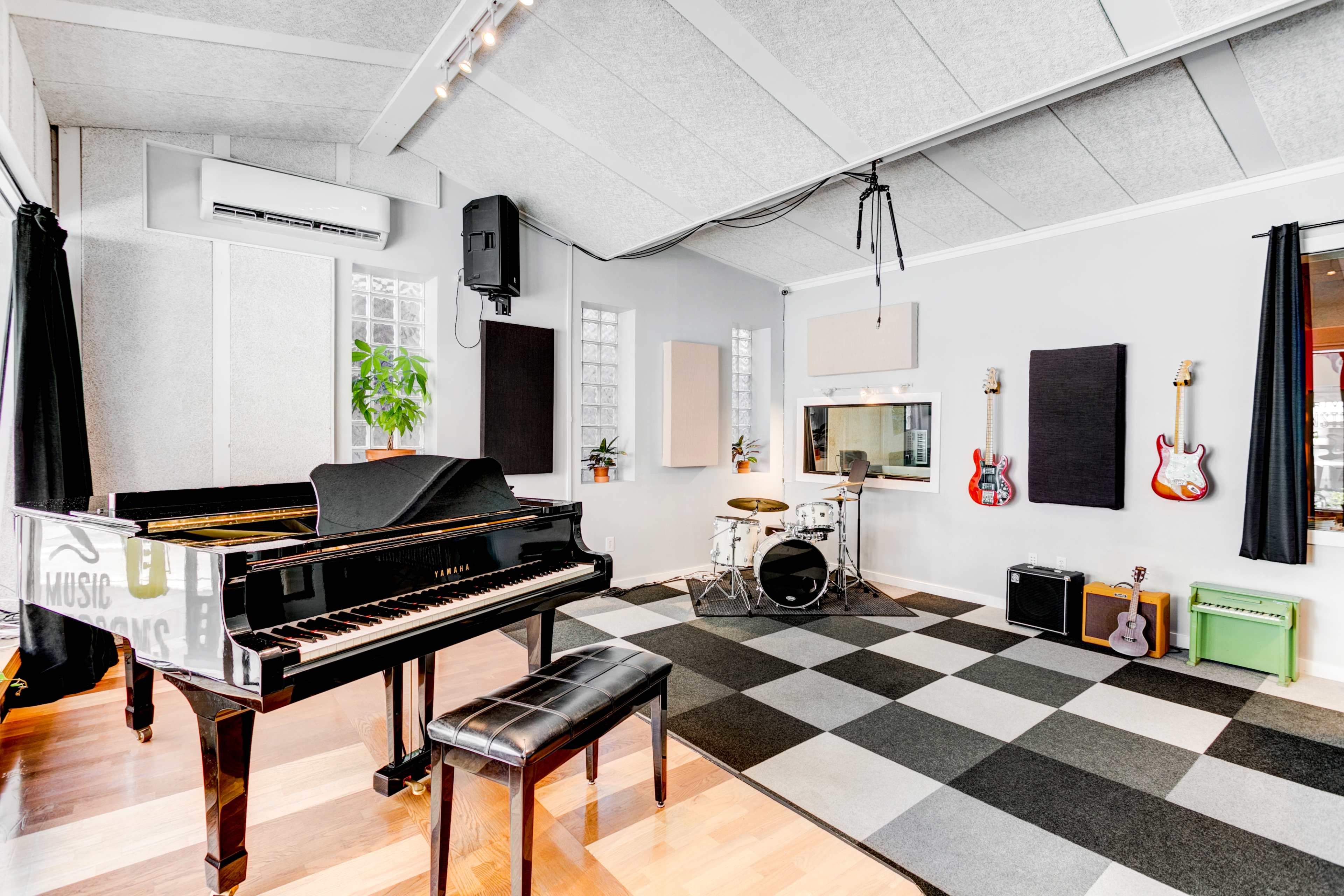 A music studio featuring a grand piano, a drum set, and various guitars displayed on the walls, with a checkerboard floor and large windows.