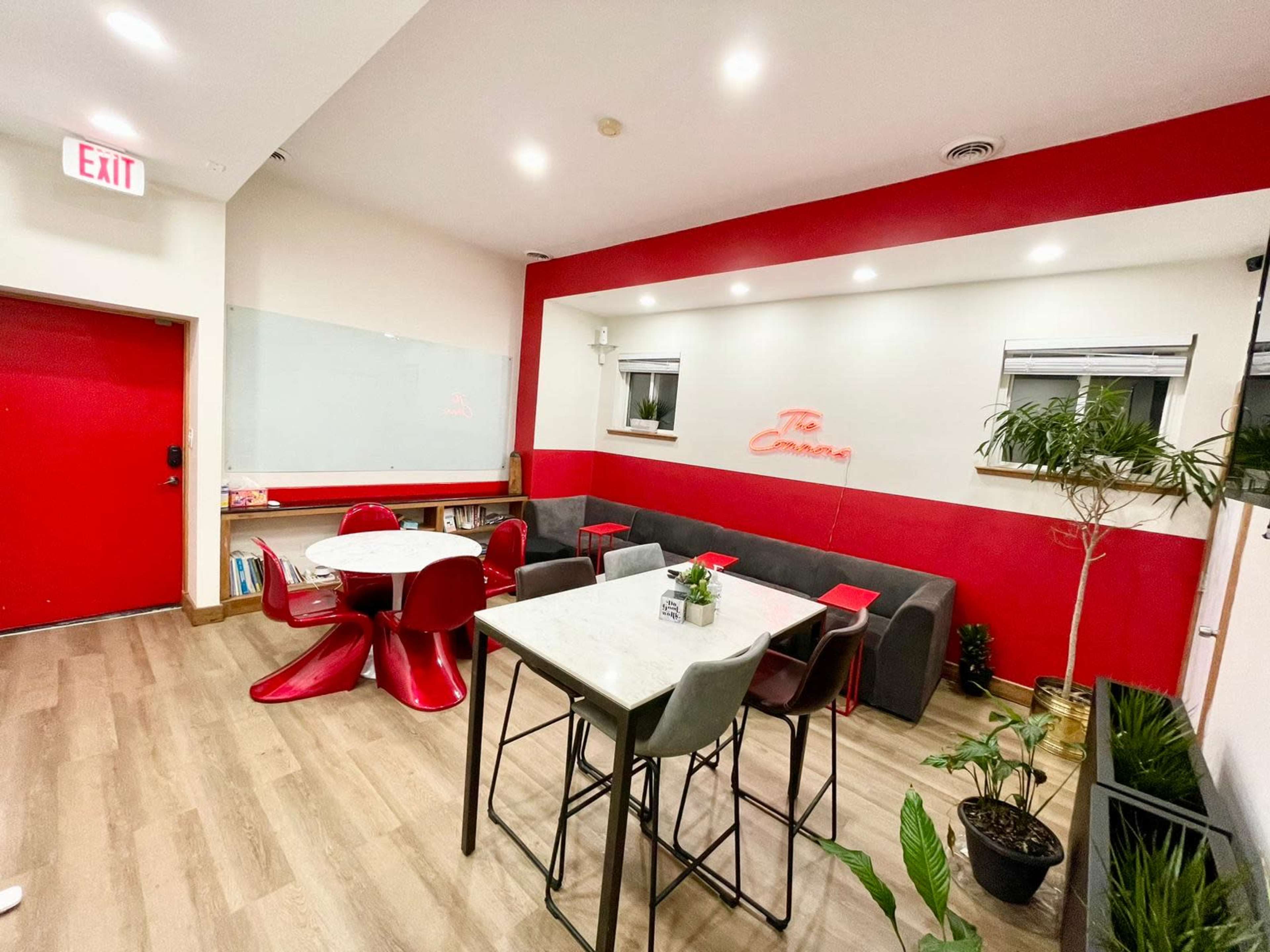 A modern, minimalist lounge area with red and white walls, featuring a few tables and chairs, along with plants in corner pots.