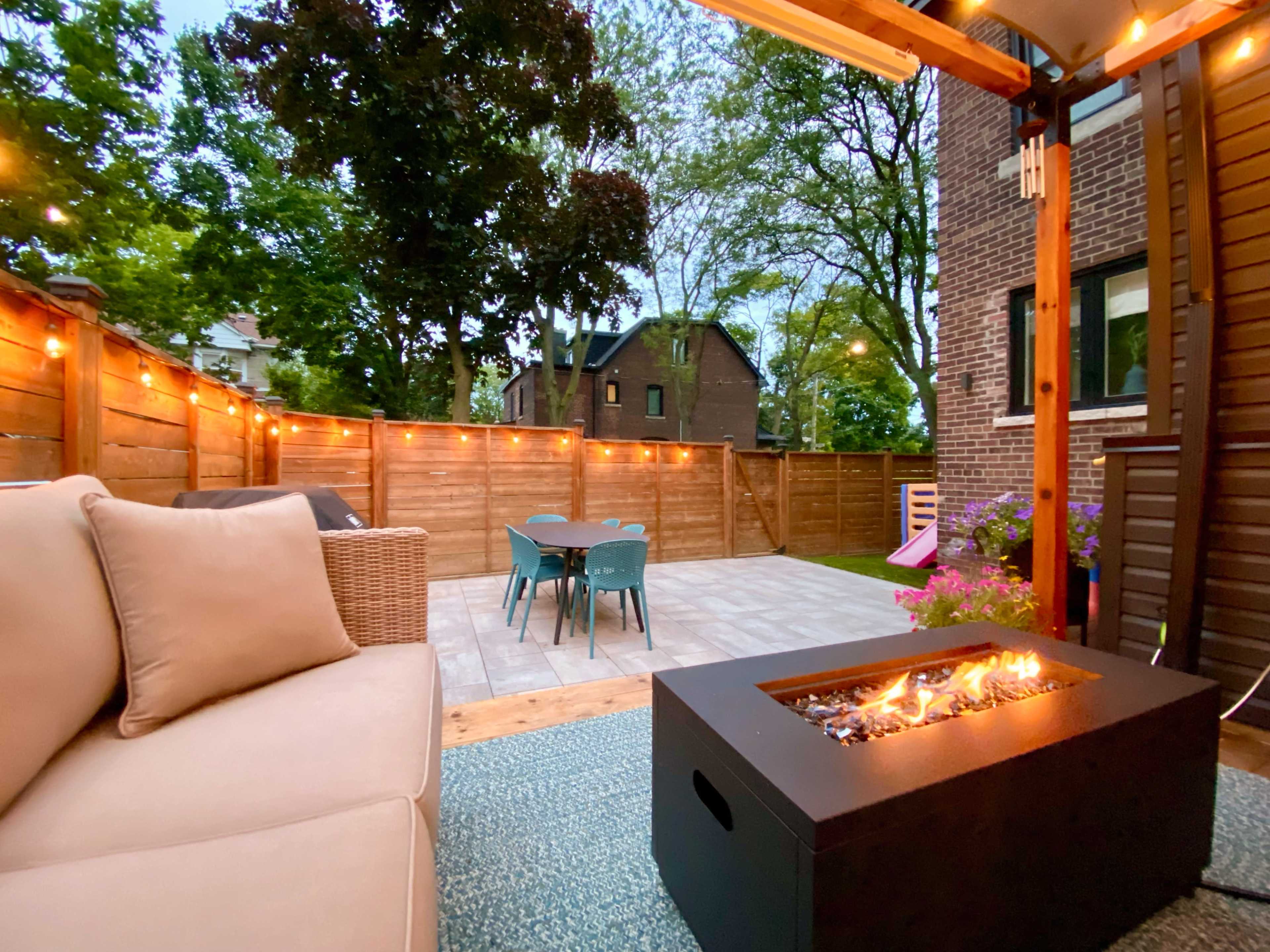The image shows a cozy outdoor patio with a fire pit, seating area, and string lights, surrounded by wooden fencing and green trees.