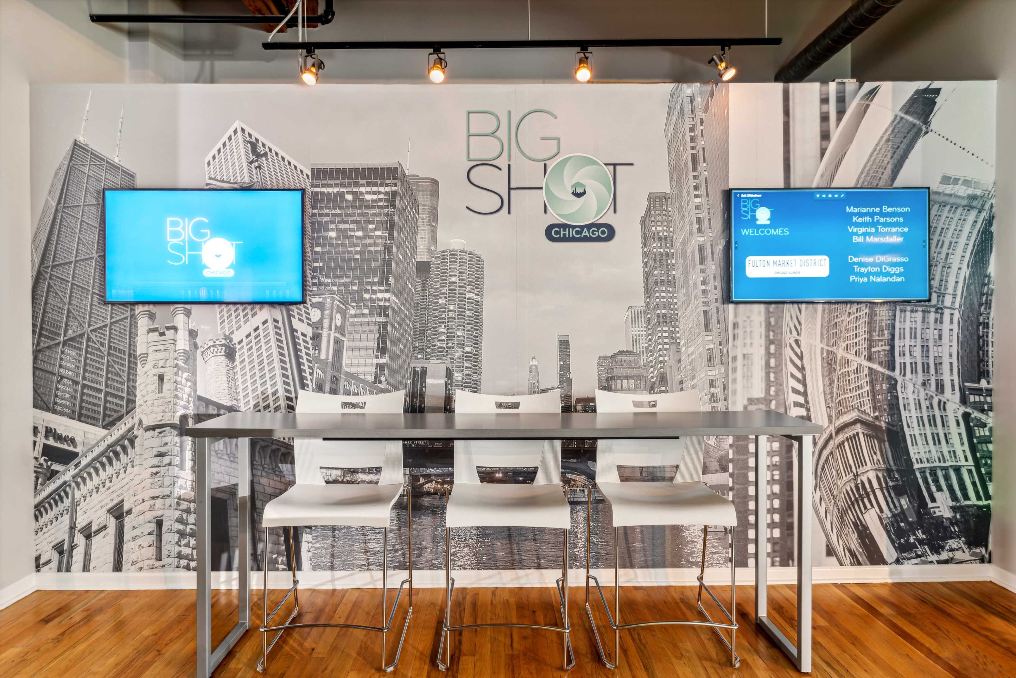 A modern meeting space featuring a table with high chairs, a large mural of Chicago's skyline, and two mounted screens displaying information.