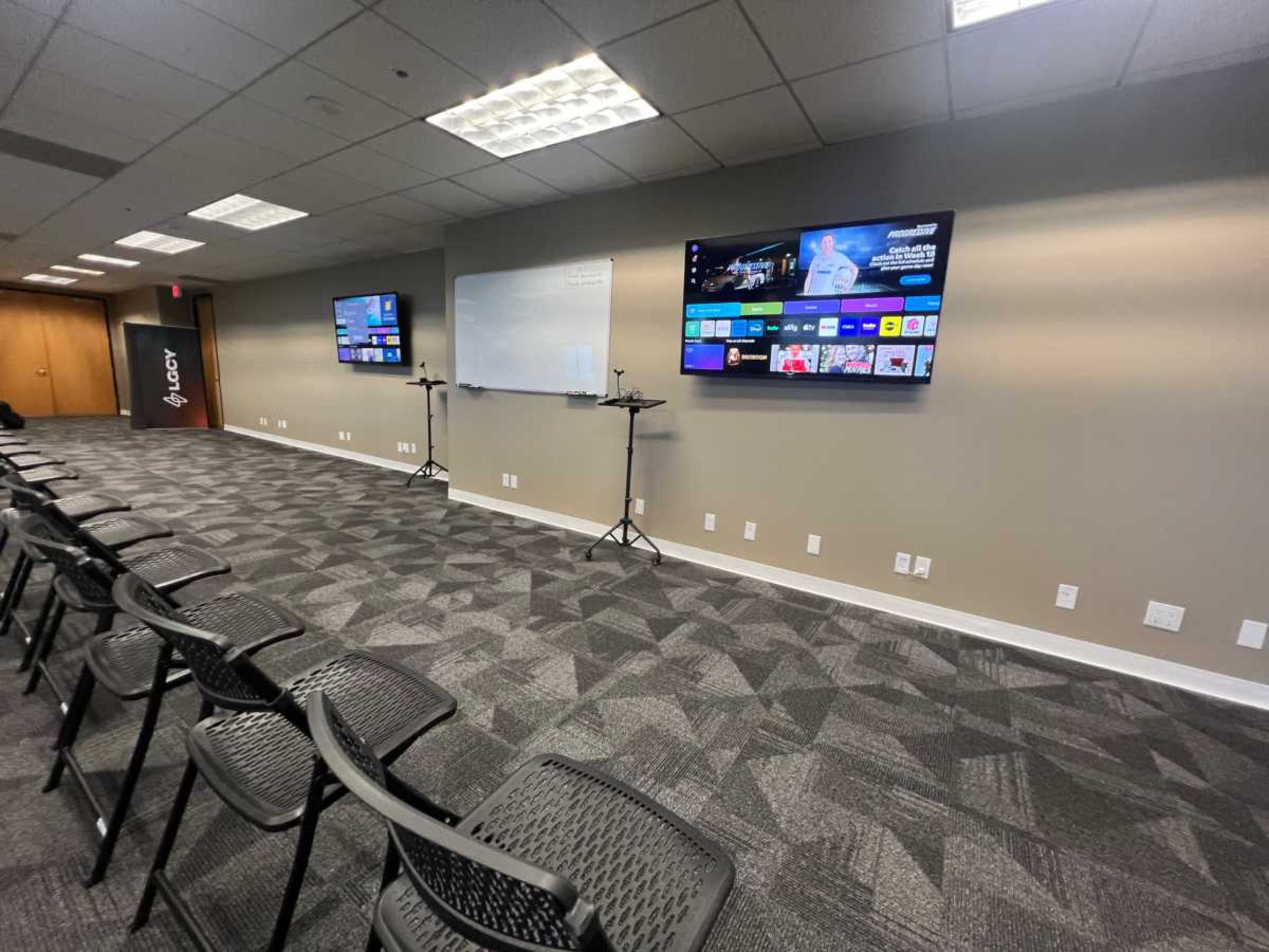 The image shows a modern meeting room with two wall-mounted screens displaying content and several black chairs arranged in rows facing the screens.
