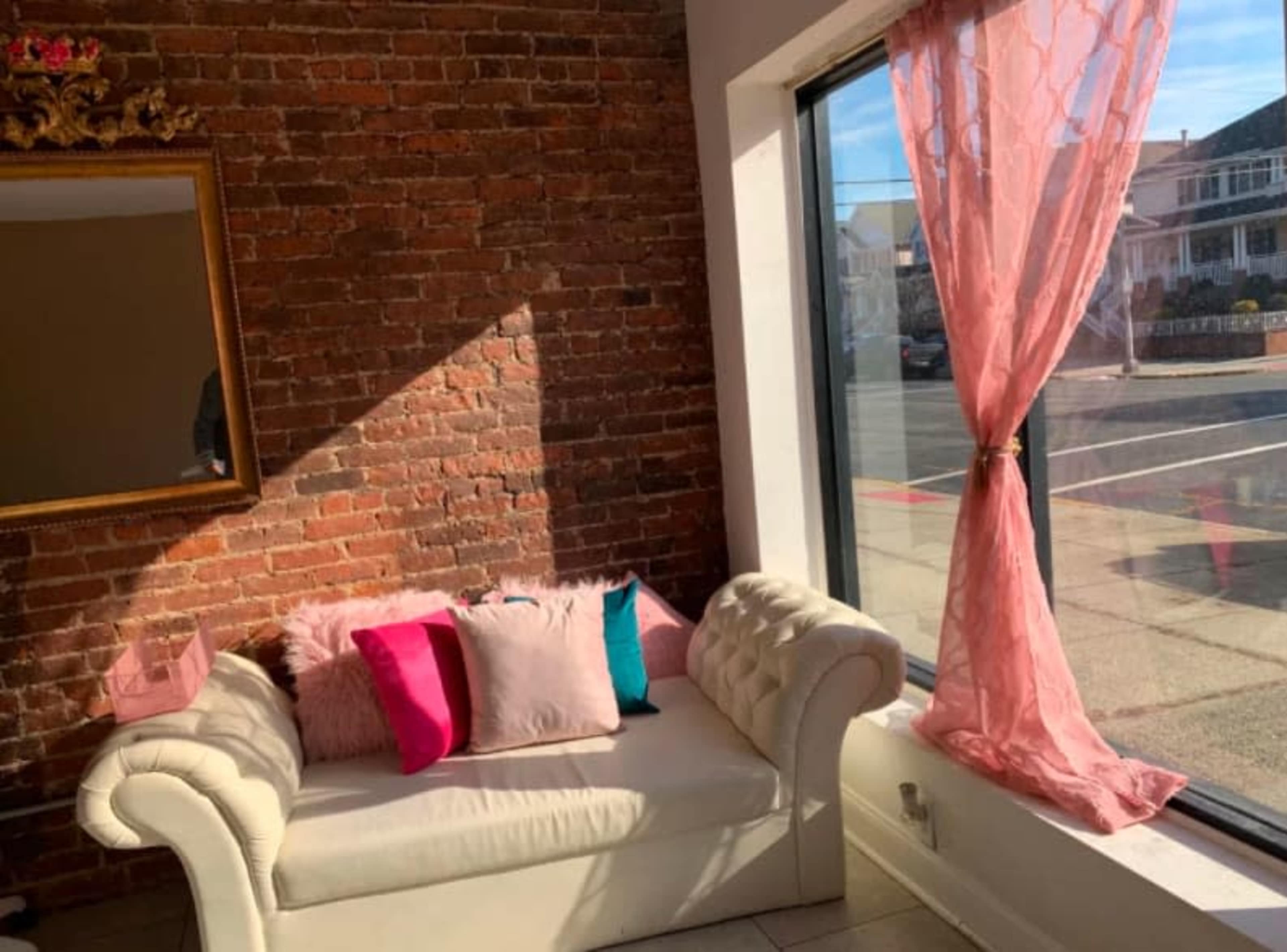 A white couch with colorful pillows positioned near a large window with pink curtains, beside a brick wall.
