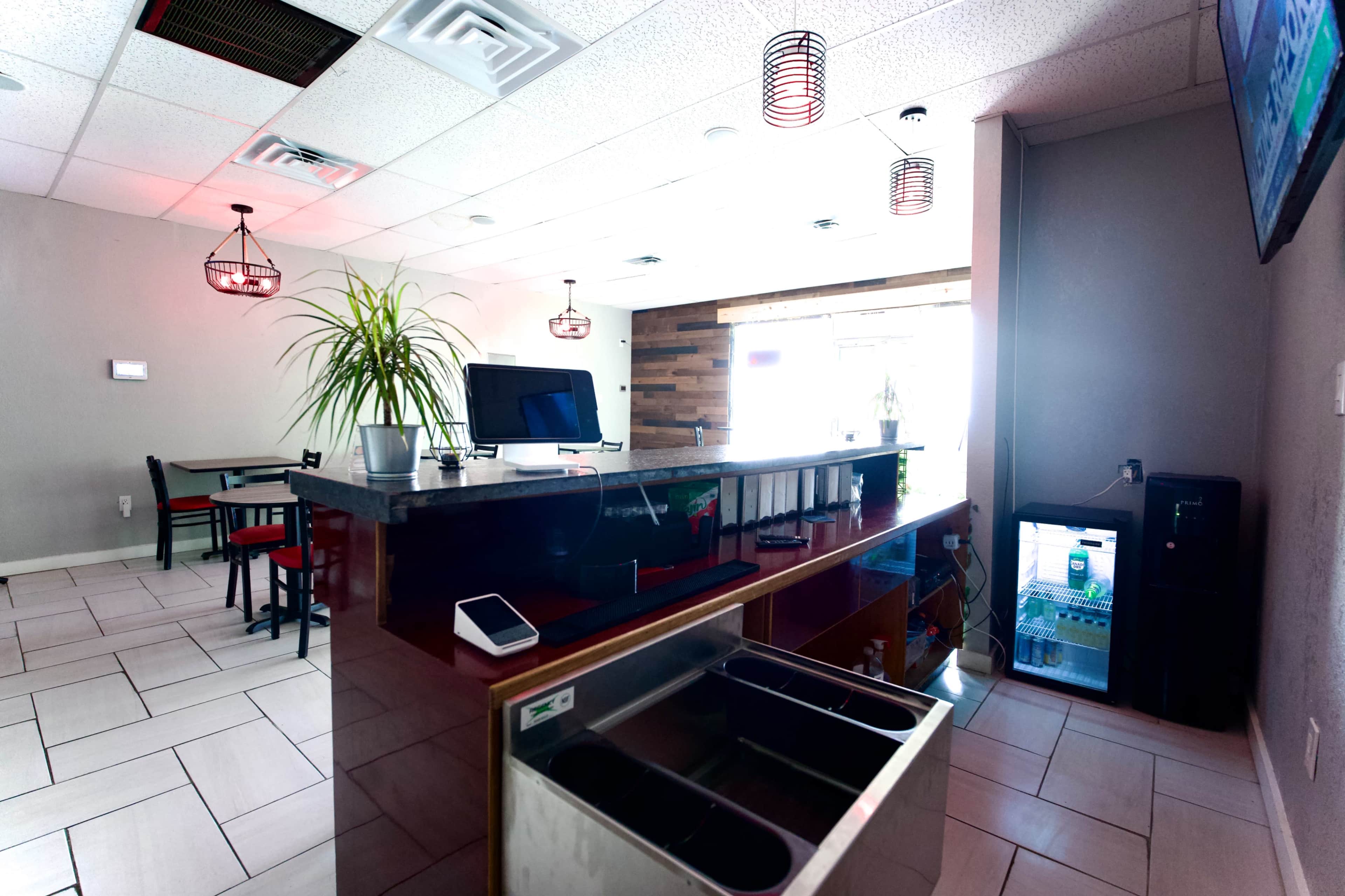 The image shows a modern interior of a restaurant or café featuring a counter with a cash register, a seating area with tables and chairs, and a display fridge in the corner.