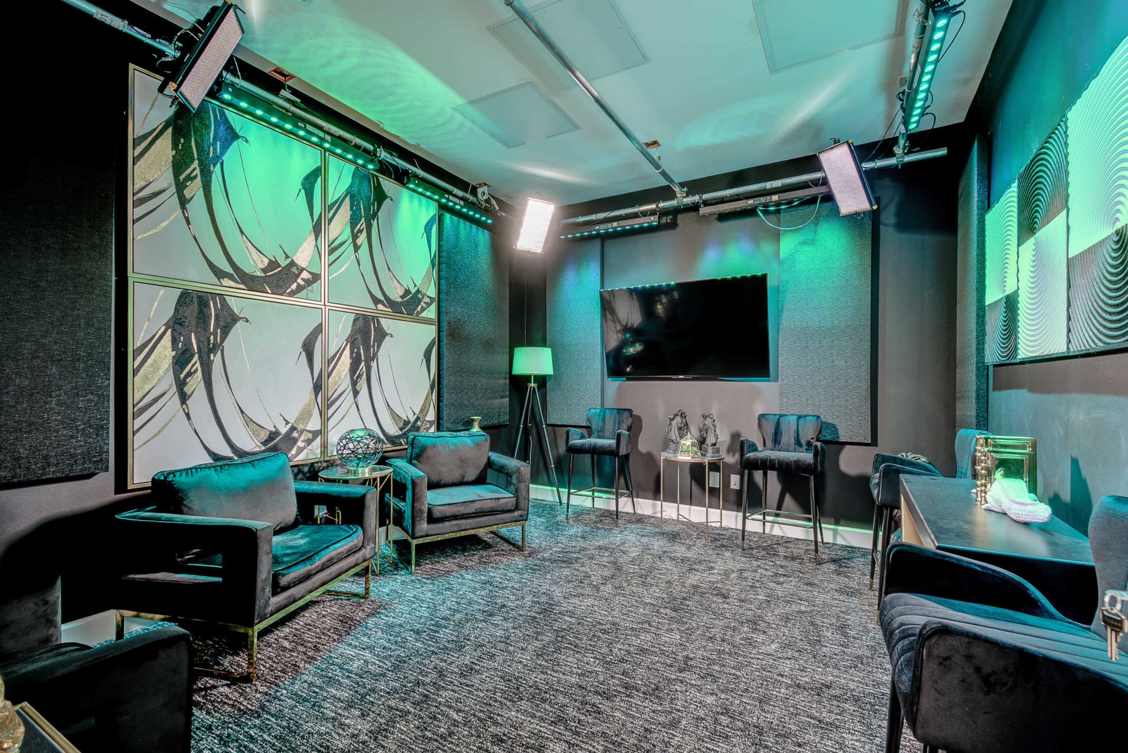 The image shows a modern studio or meeting space with dark furnishings, a large screen, and ambient green lighting.