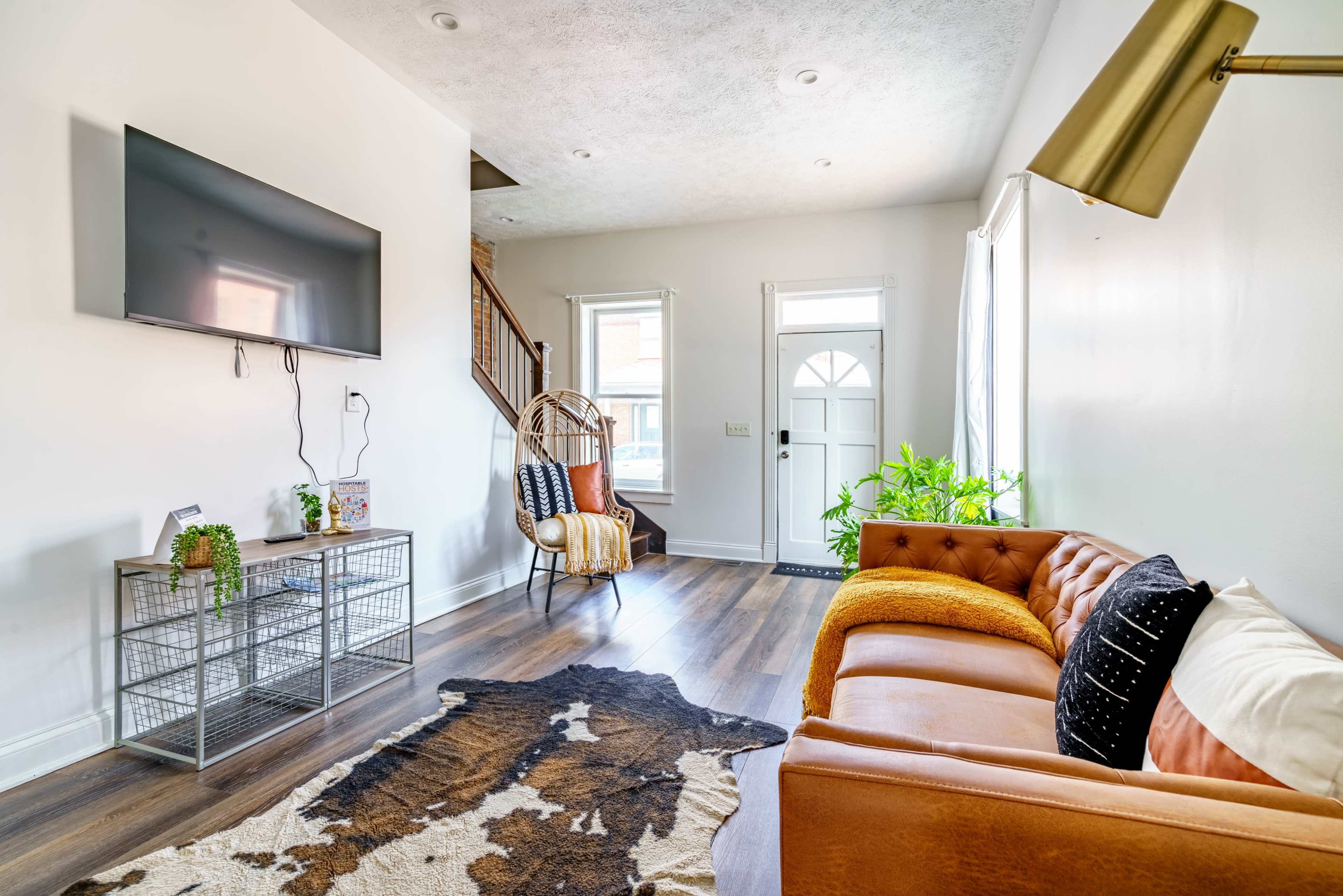 The image shows a modern living room with a brown leather sofa, a television mounted on the wall, and a plant next to a staircase.