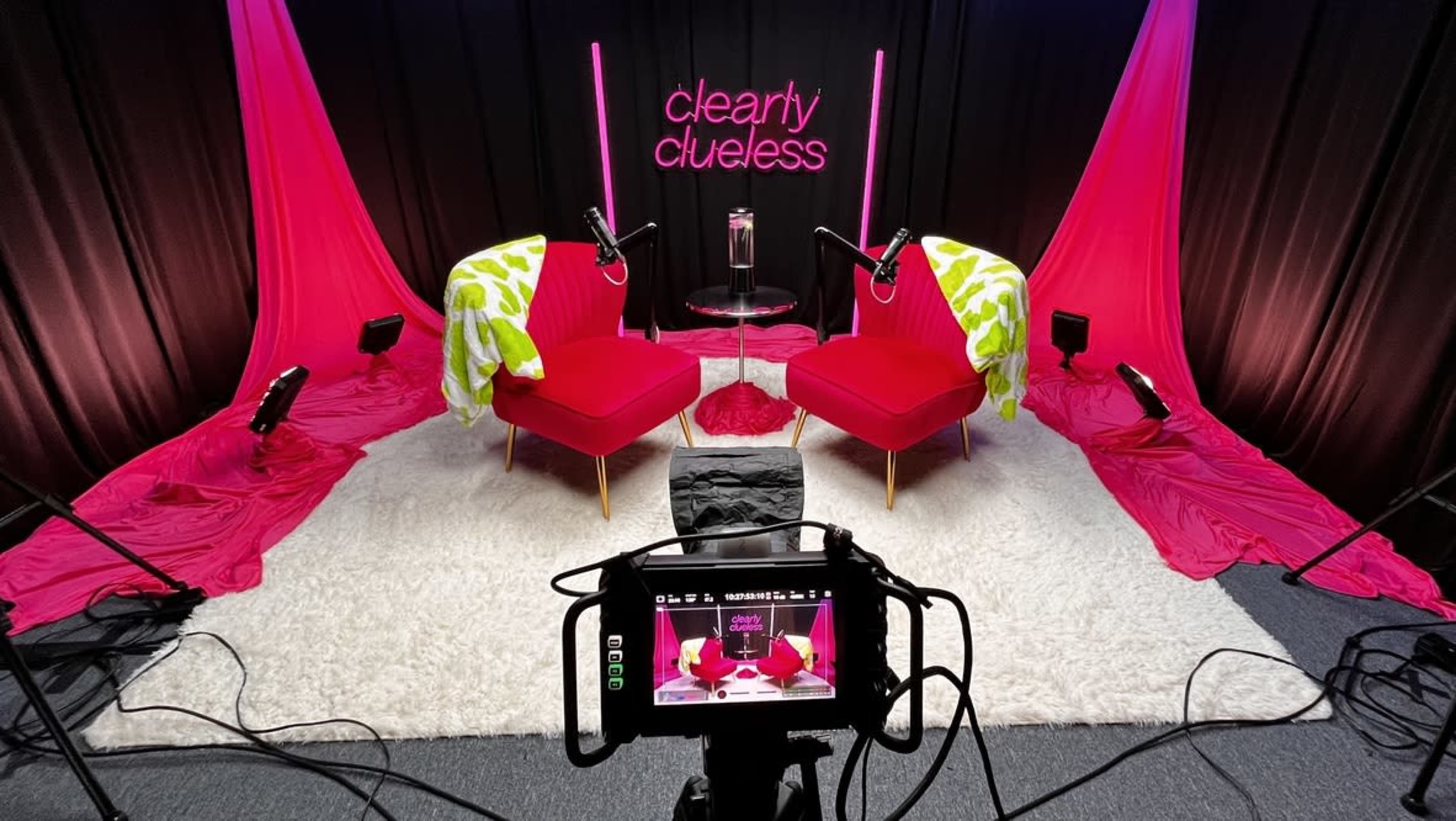 The image shows a vibrant studio setup with two red chairs, a small glass table, and bright pink fabrics draped in the background, all illuminated by spotlights.