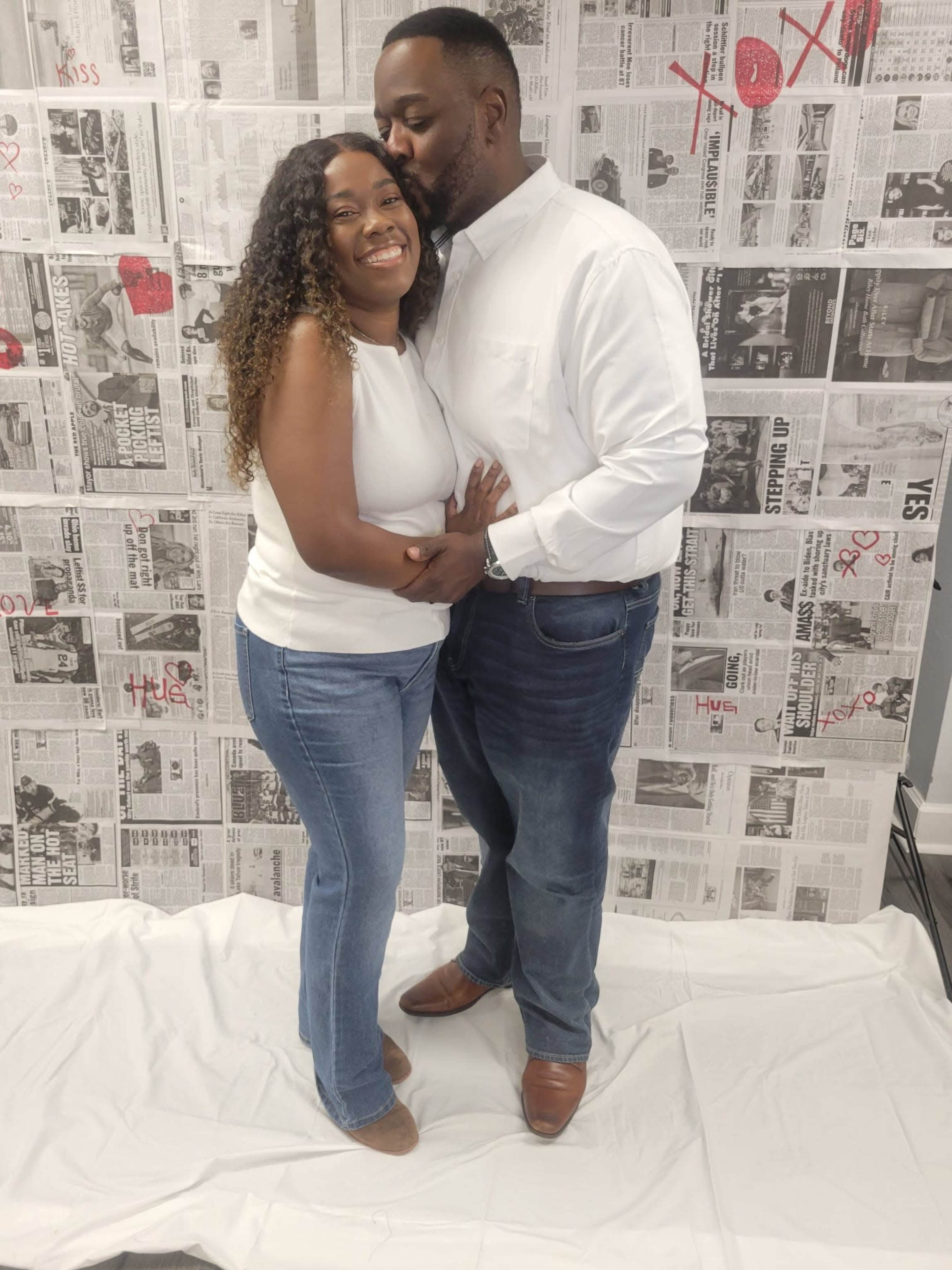 A couple stands together in front of a newspaper-themed backdrop, with the man leaning in to kiss the woman on the cheek.
