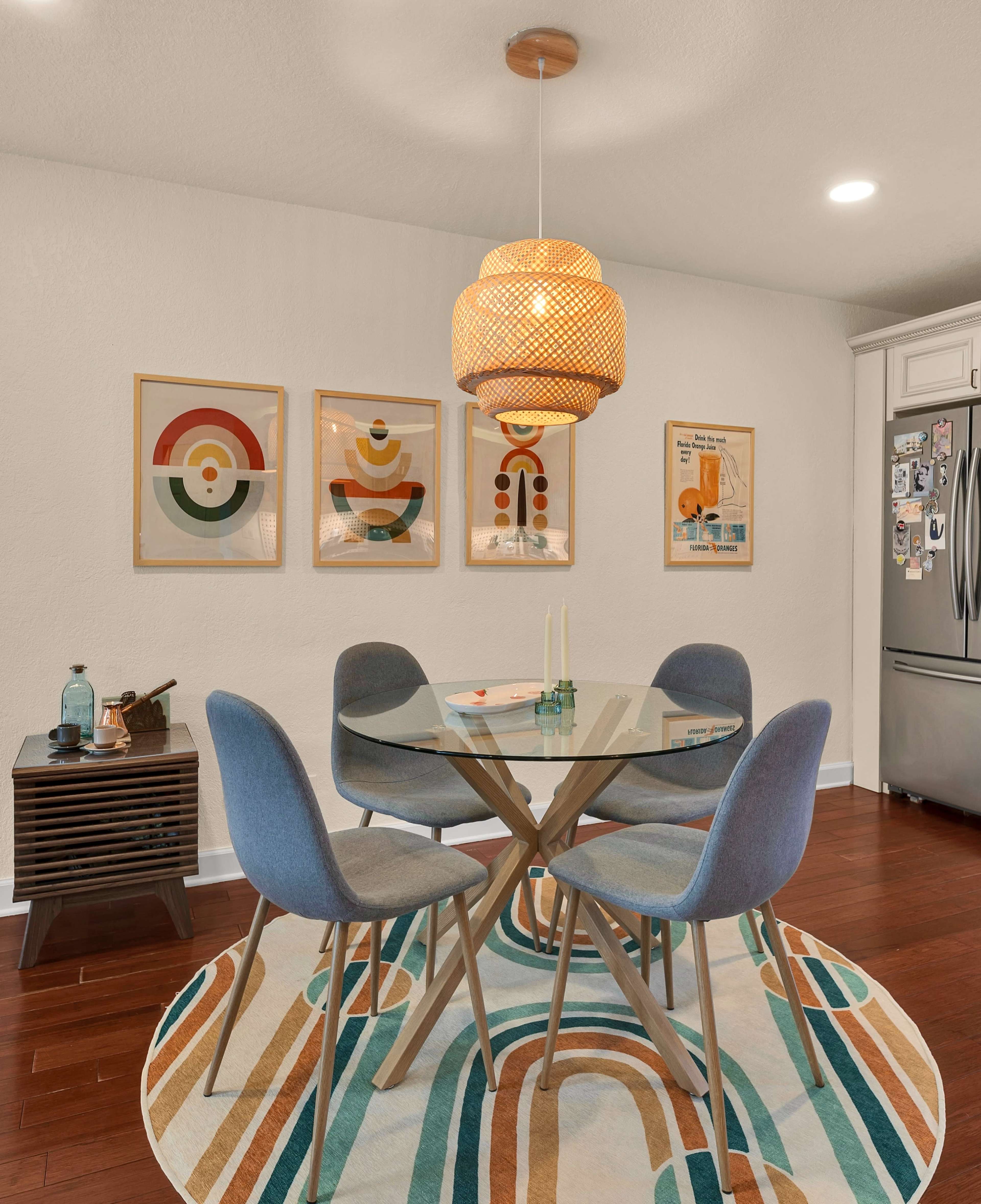 A dining area features a round glass table surrounded by four blue chairs, a patterned area rug, and framed artworks on the wall above.
