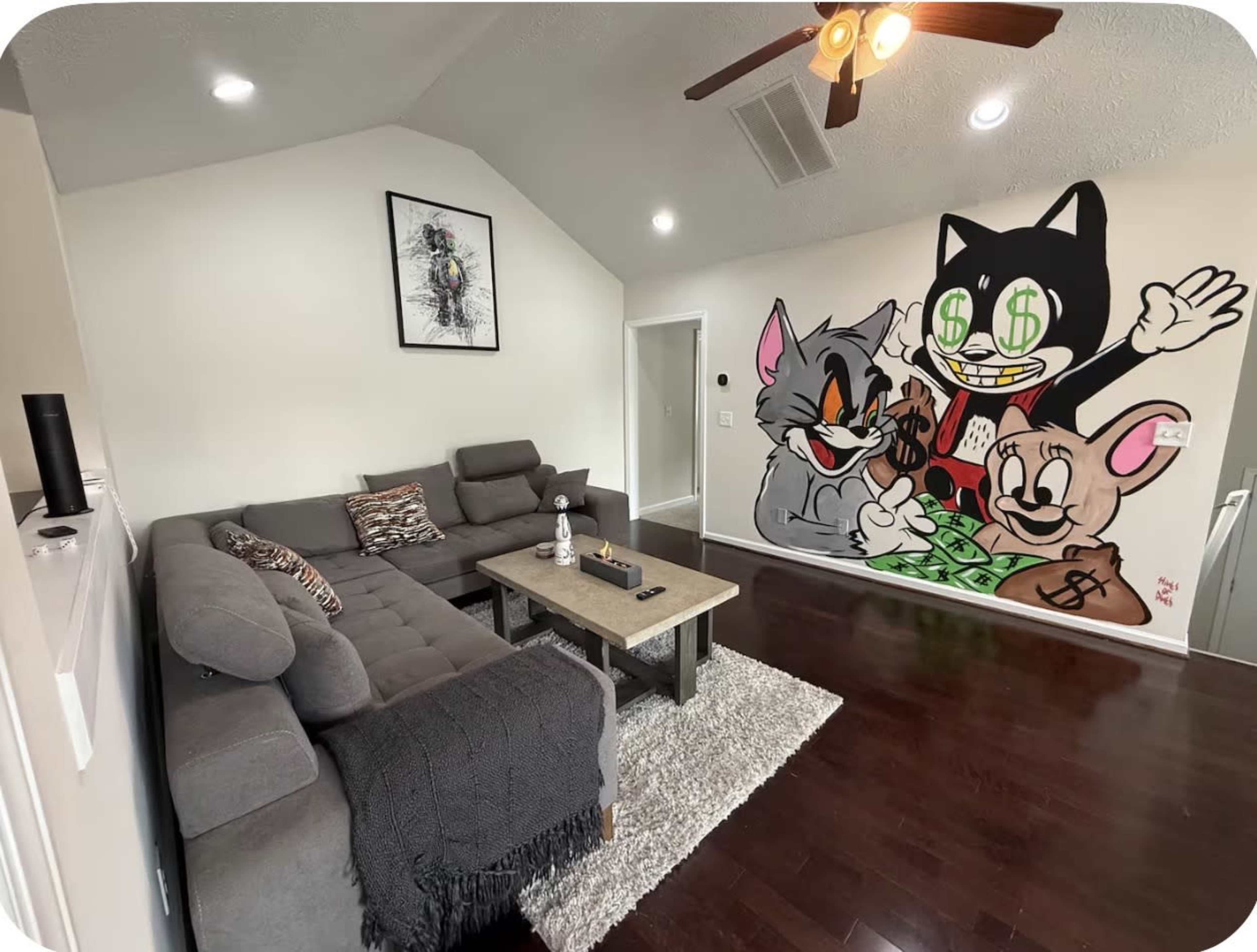 The image shows a living room with a gray sectional sofa, a wooden coffee table, and a large mural featuring cartoon characters on one wall.