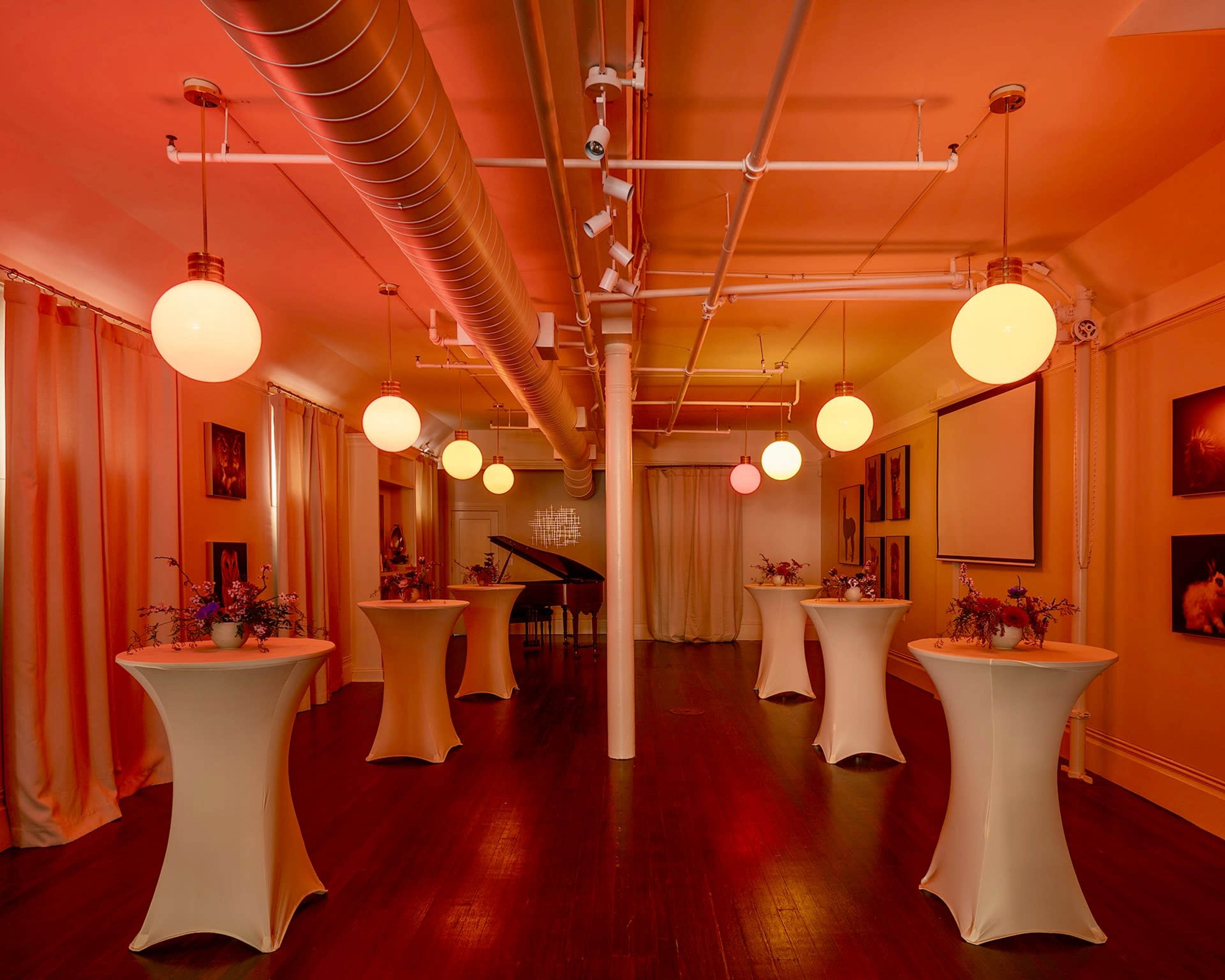 A spacious room with tall ceilings, featuring several round tables covered with white cloths and illuminated by large pendant lights.
