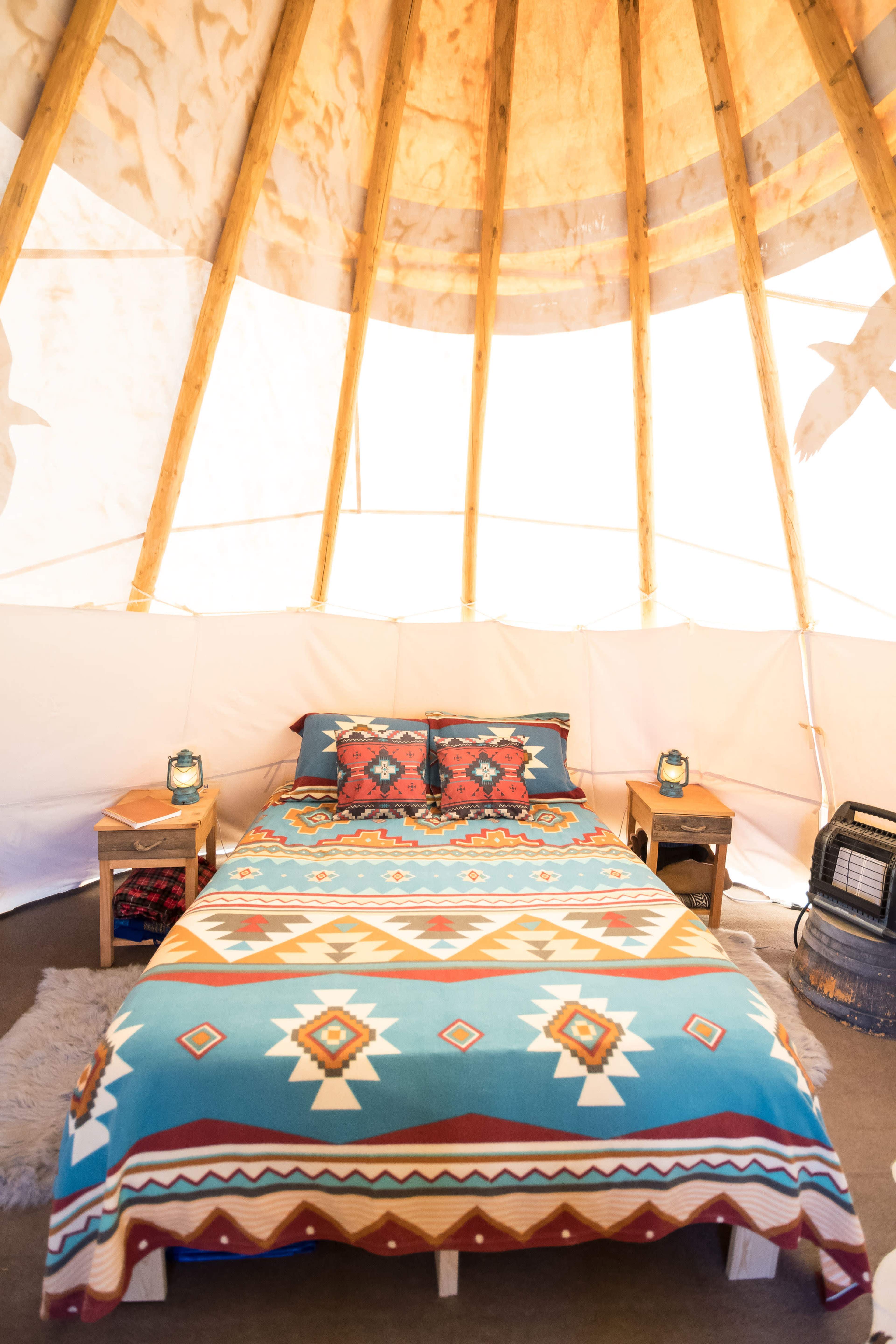 The interior of a tip tent features a bed with a colorful patterned blanket and two nightstands with lanterns.