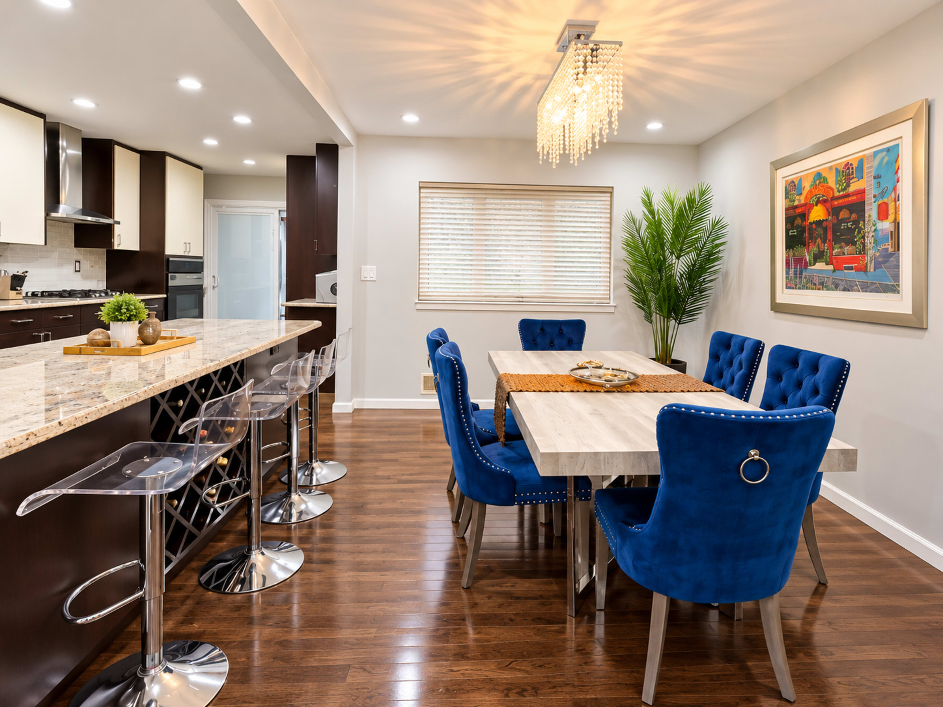 The image shows a modern dining area featuring a wooden table with blue velvet chairs, a decorative chandelier, and a kitchen bar with clear stools.