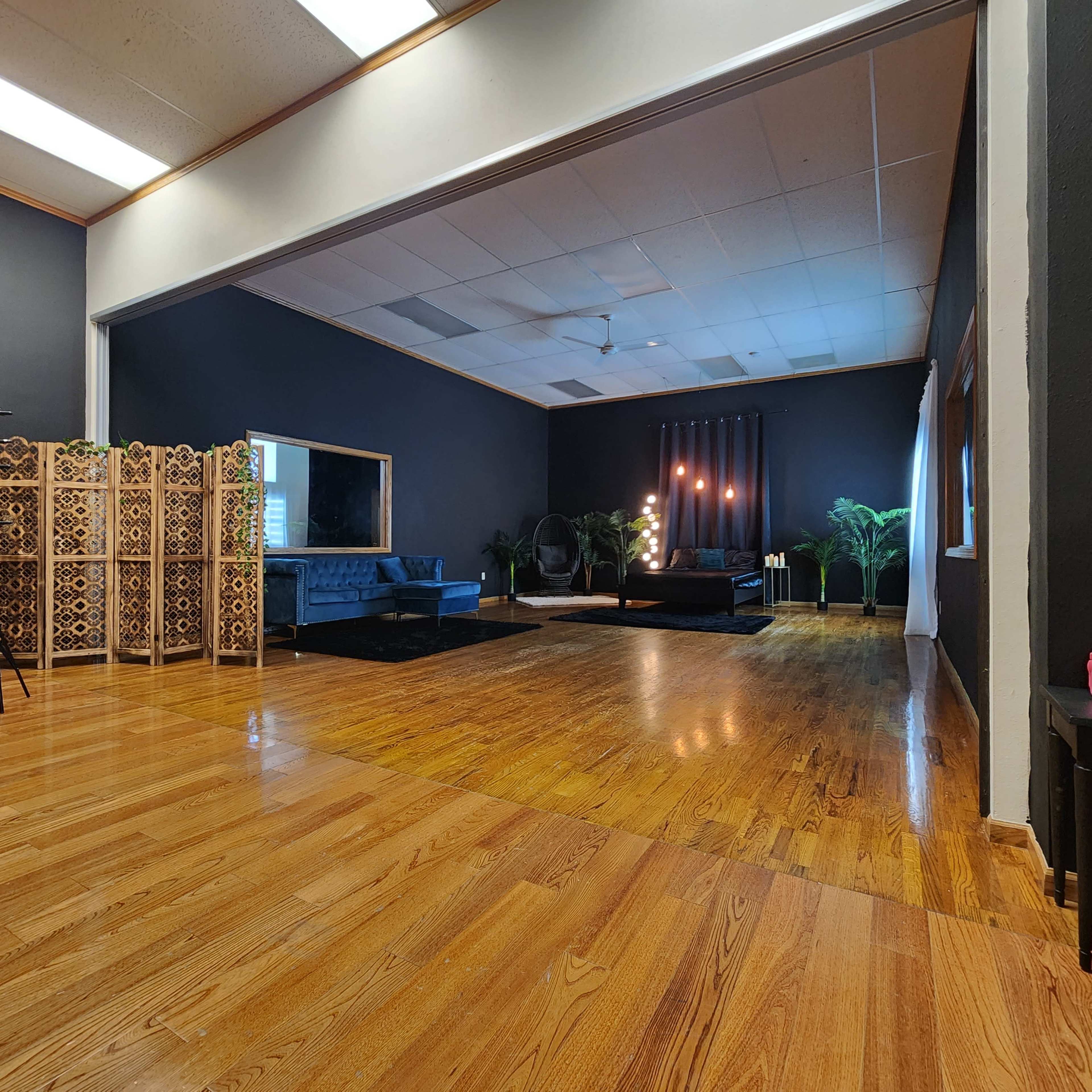 A spacious room with wooden flooring, featuring a dark wall, a blue sofa, decorative plants, and a partition made of intricate wooden panels.