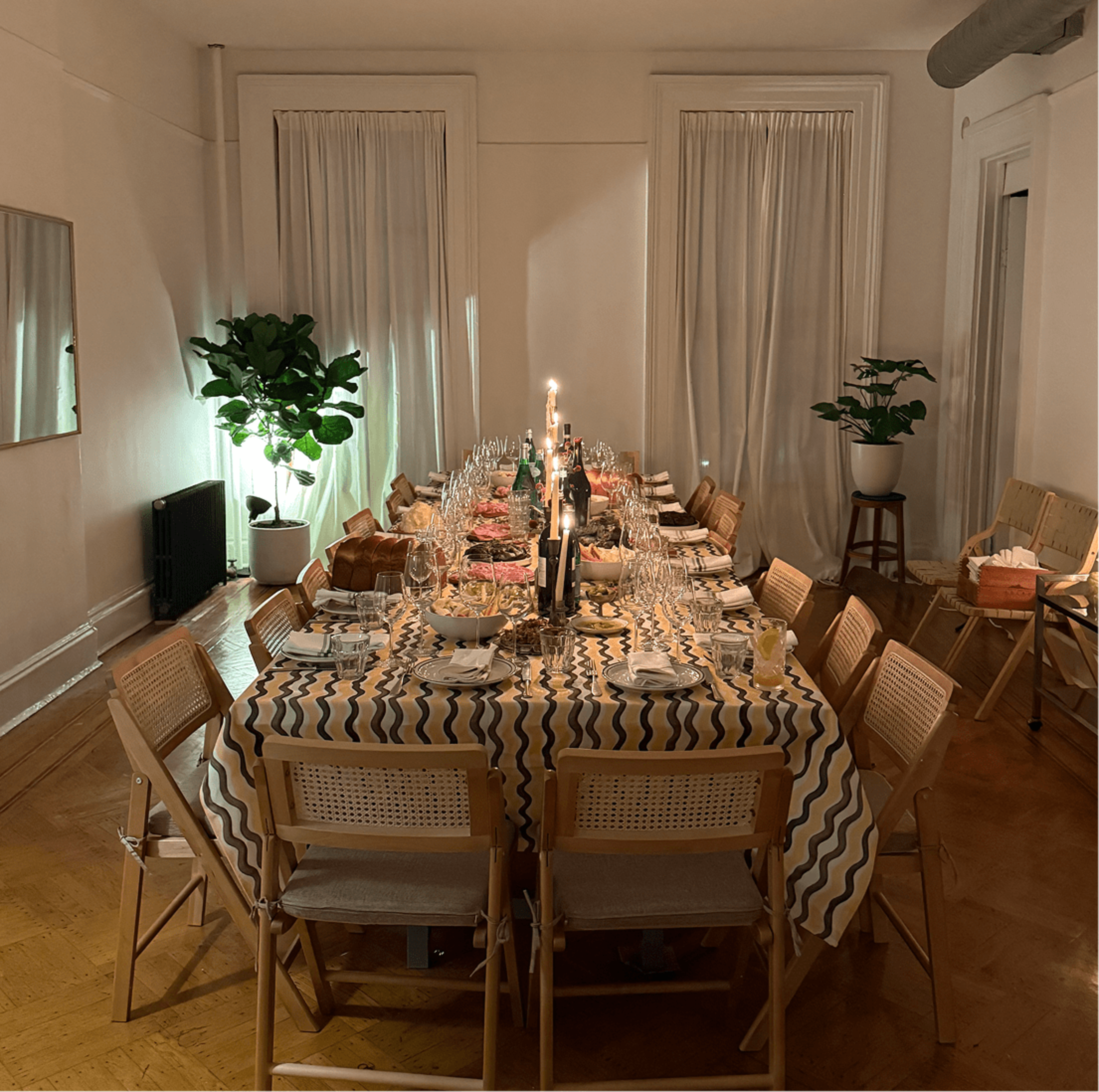 A long dining table is set with plates and glasses, surrounded by wooden chairs in a warmly lit room.