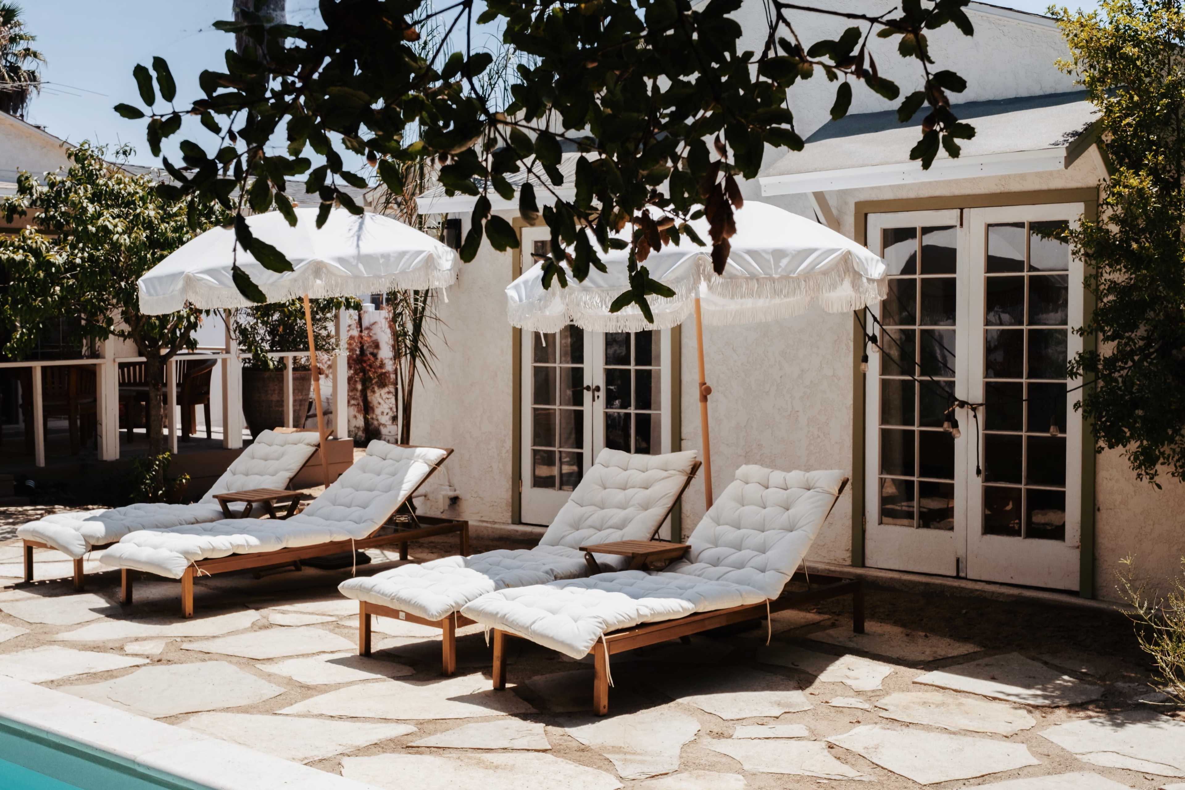 A set of four lounge chairs with white cushions and umbrellas is positioned next to a swimming pool beside a house with French doors.