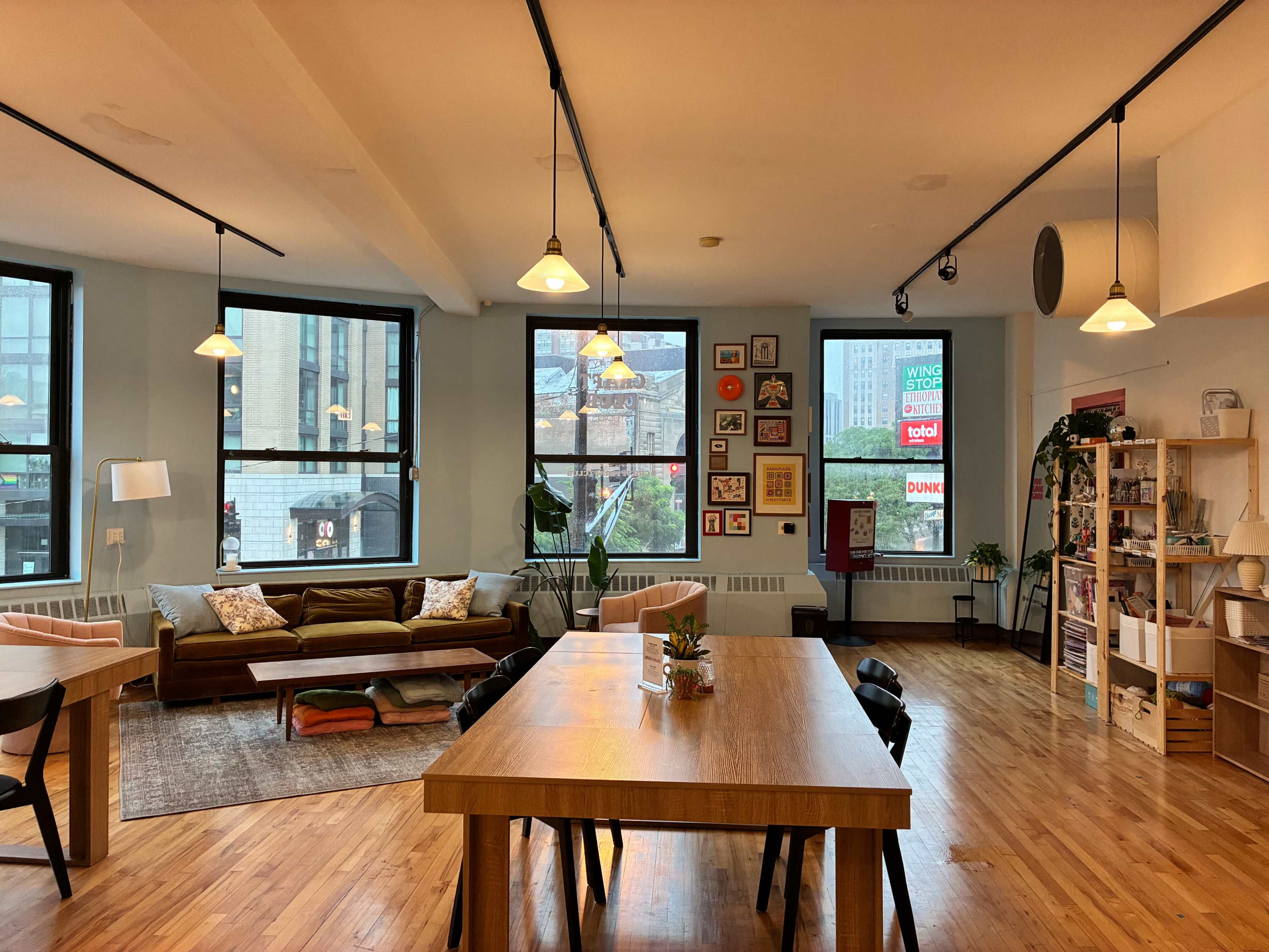 The image shows a modern co-working space with large windows, wooden flooring, and a mix of seating areas that include a dining table and a cozy lounge area.