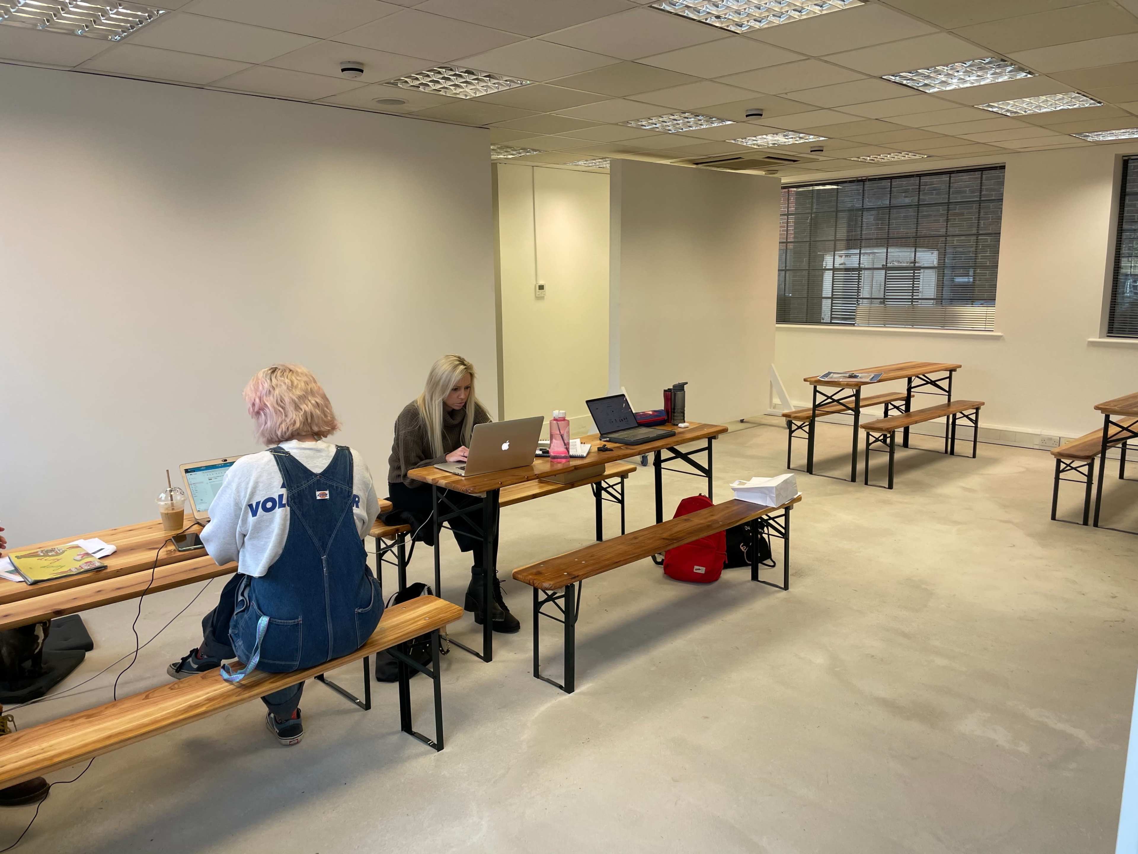 Two people work on laptops at wooden tables in a sparsely furnished room with large windows.