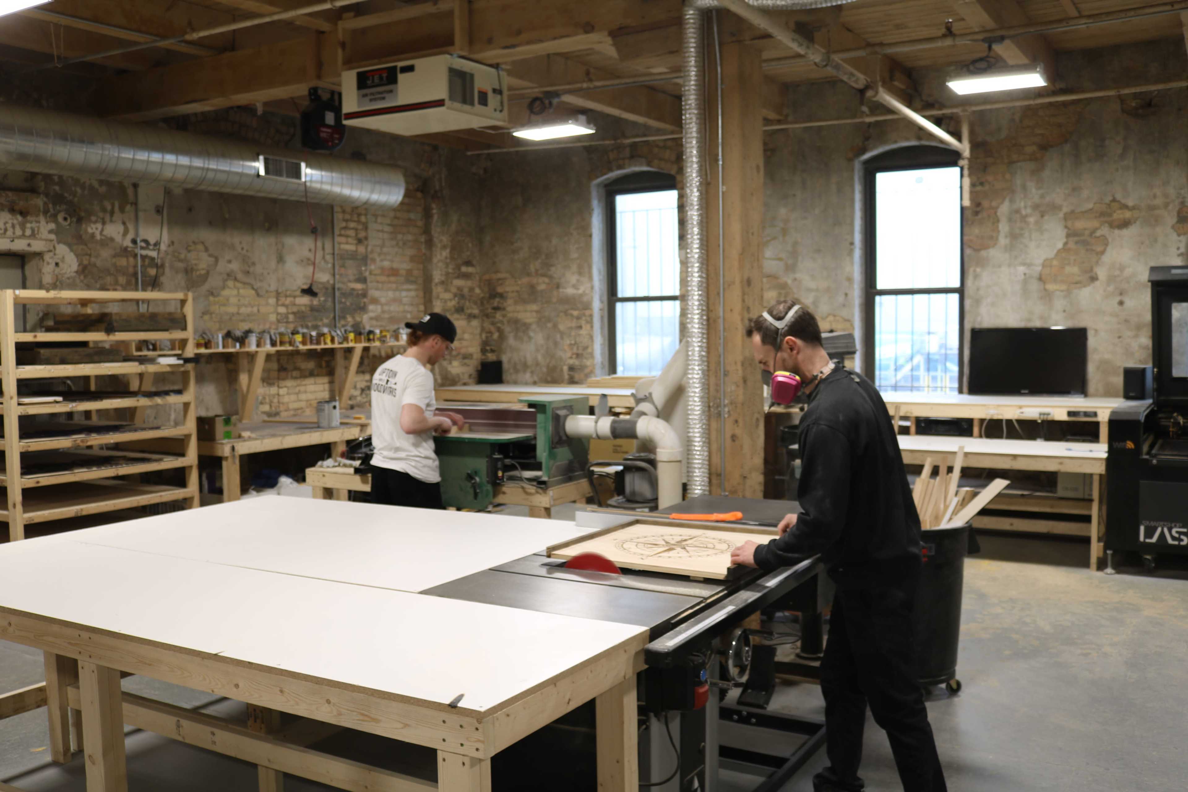 Two individuals are working in a woodshop, one operating a table saw and the other using a planer.