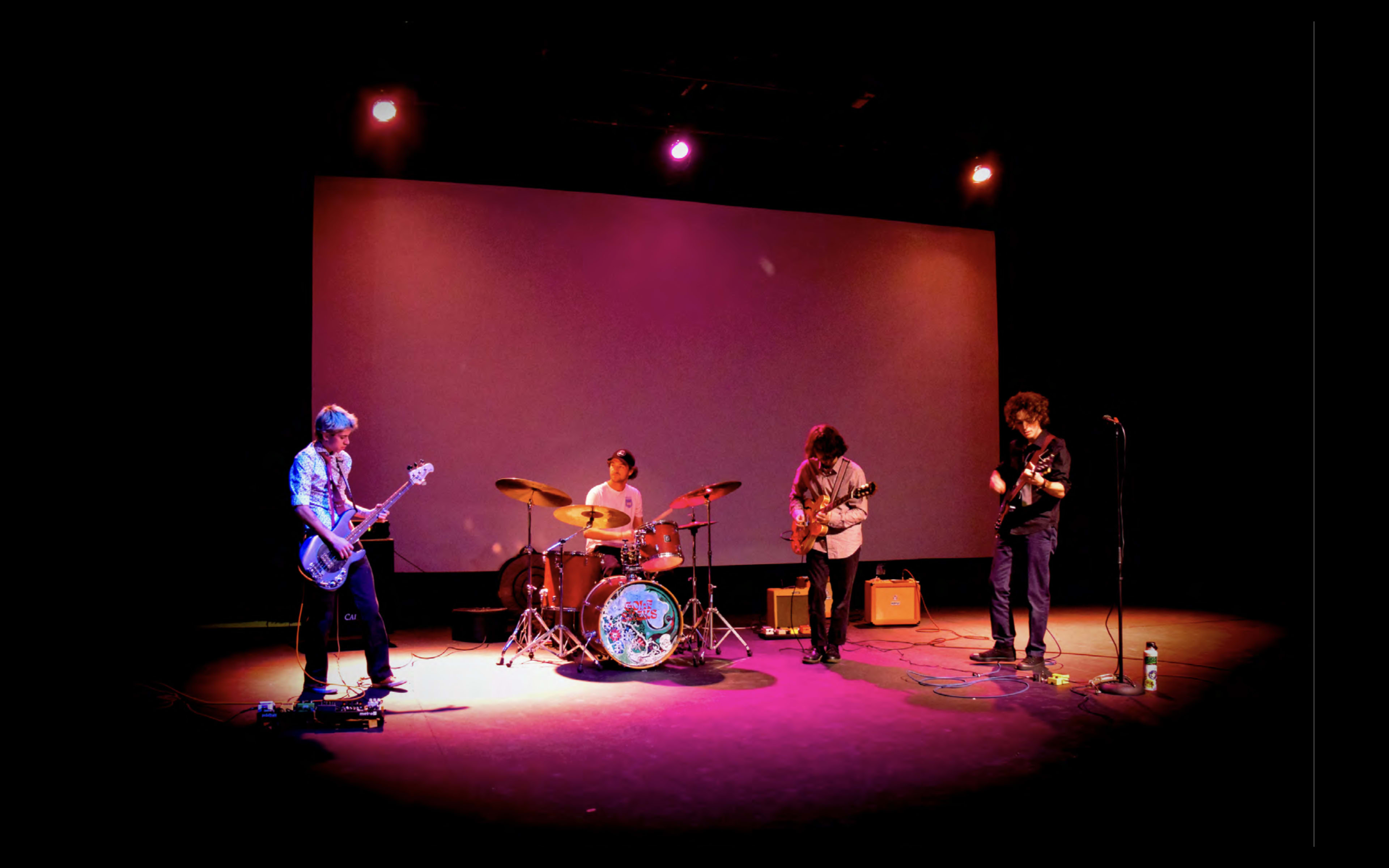 A four-member band performs on a dimly lit stage with a large blank screen in the background.