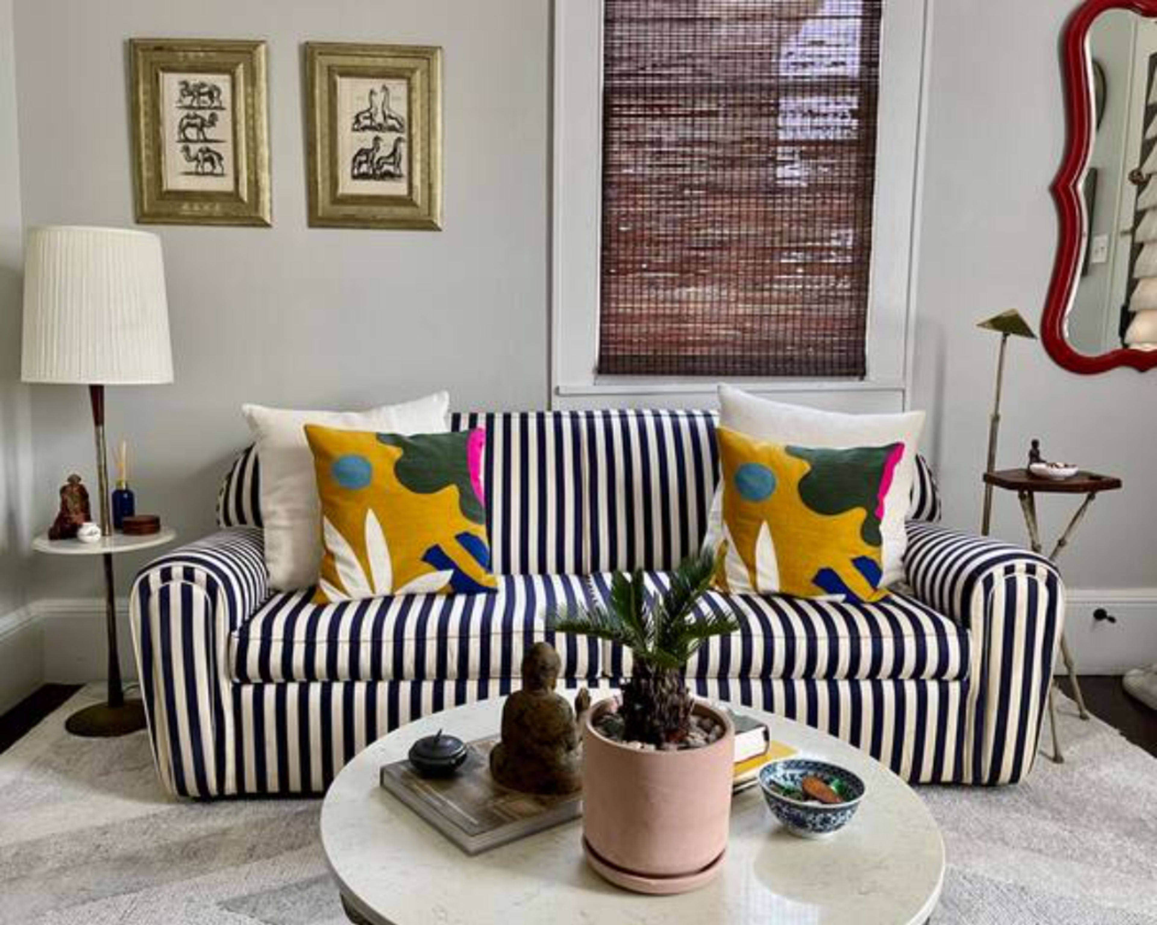 A living room featuring a striped sofa with colorful pillows, a round coffee table with decorative items, and a window with bamboo shades.