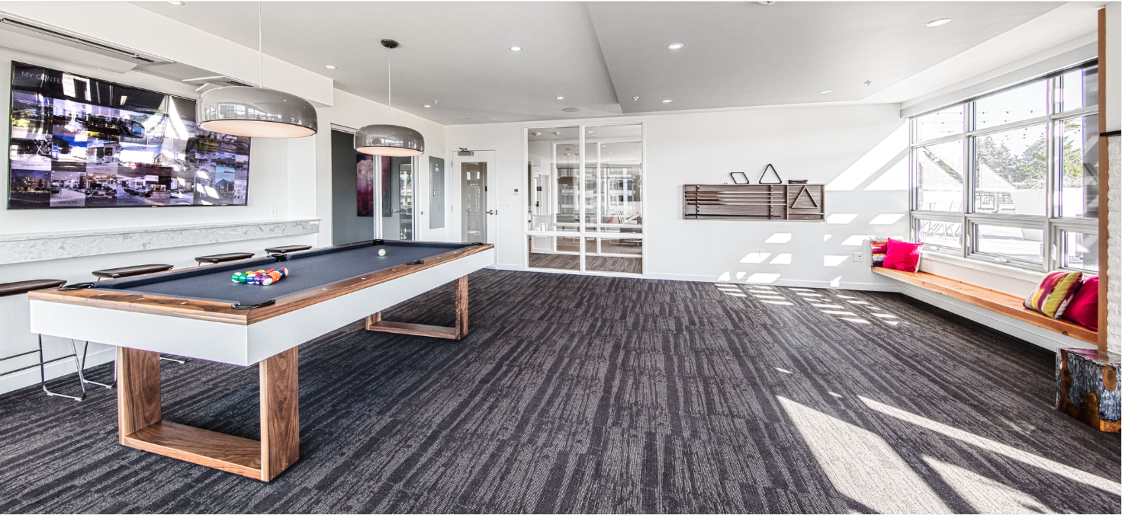 The image shows a modern lounge area featuring a black pool table, wooden bench seating, and large windows allowing natural light to fill the space.
