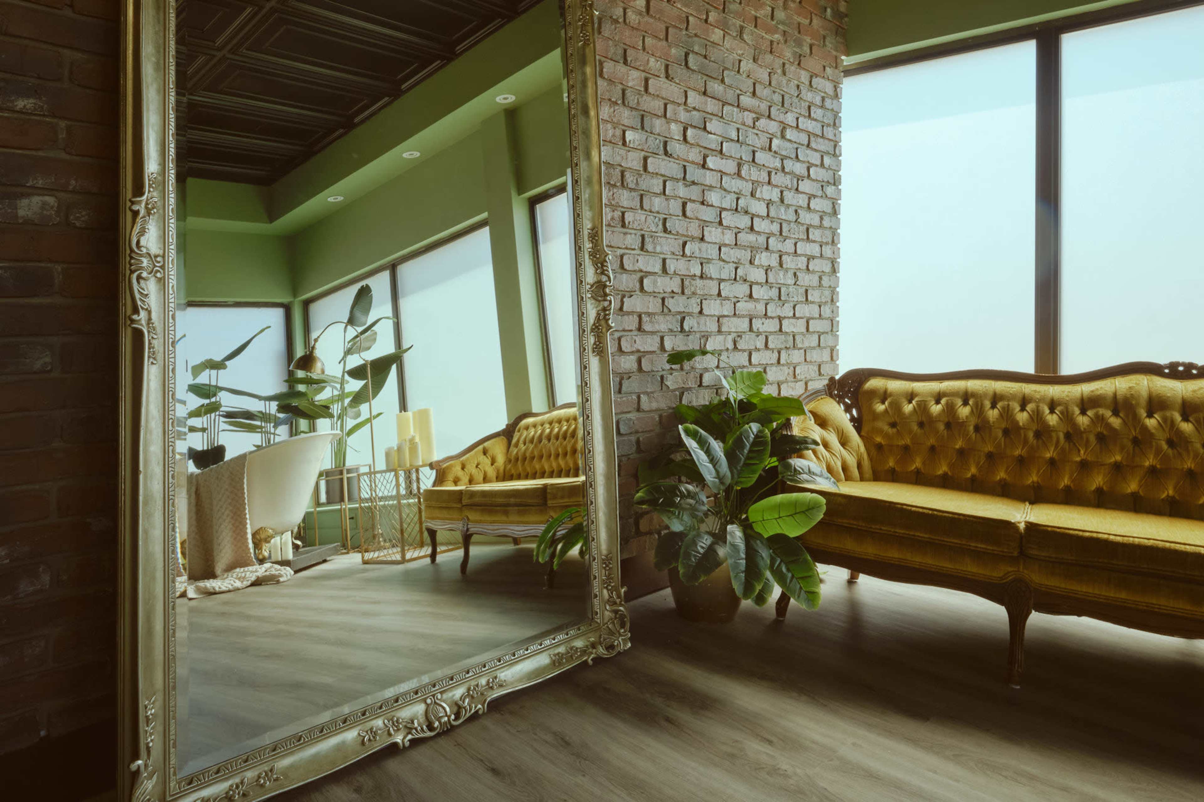 The image features a spacious room with a large ornate mirror reflecting a vintage yellow sofa and potted plants against a brick wall.