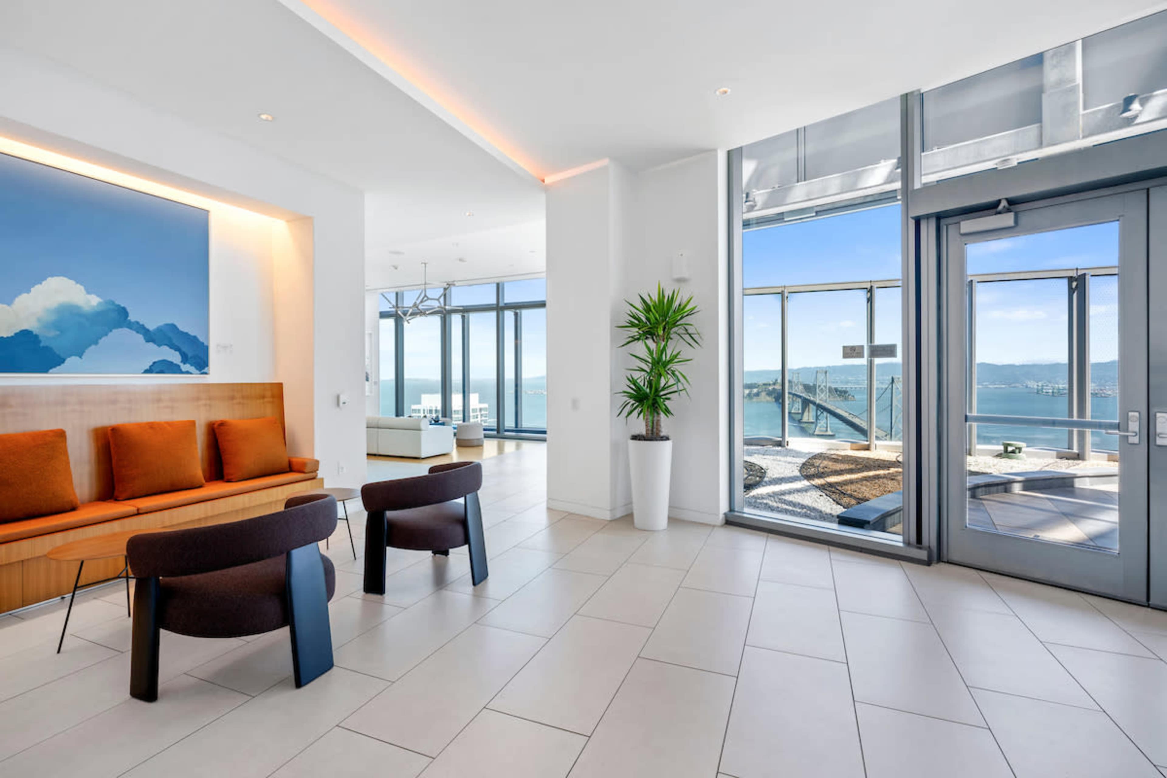 The image shows a modern lounge area with a sofa and two chairs, large windows offering a view of a bridge and water, and a potted plant near the entrance.
