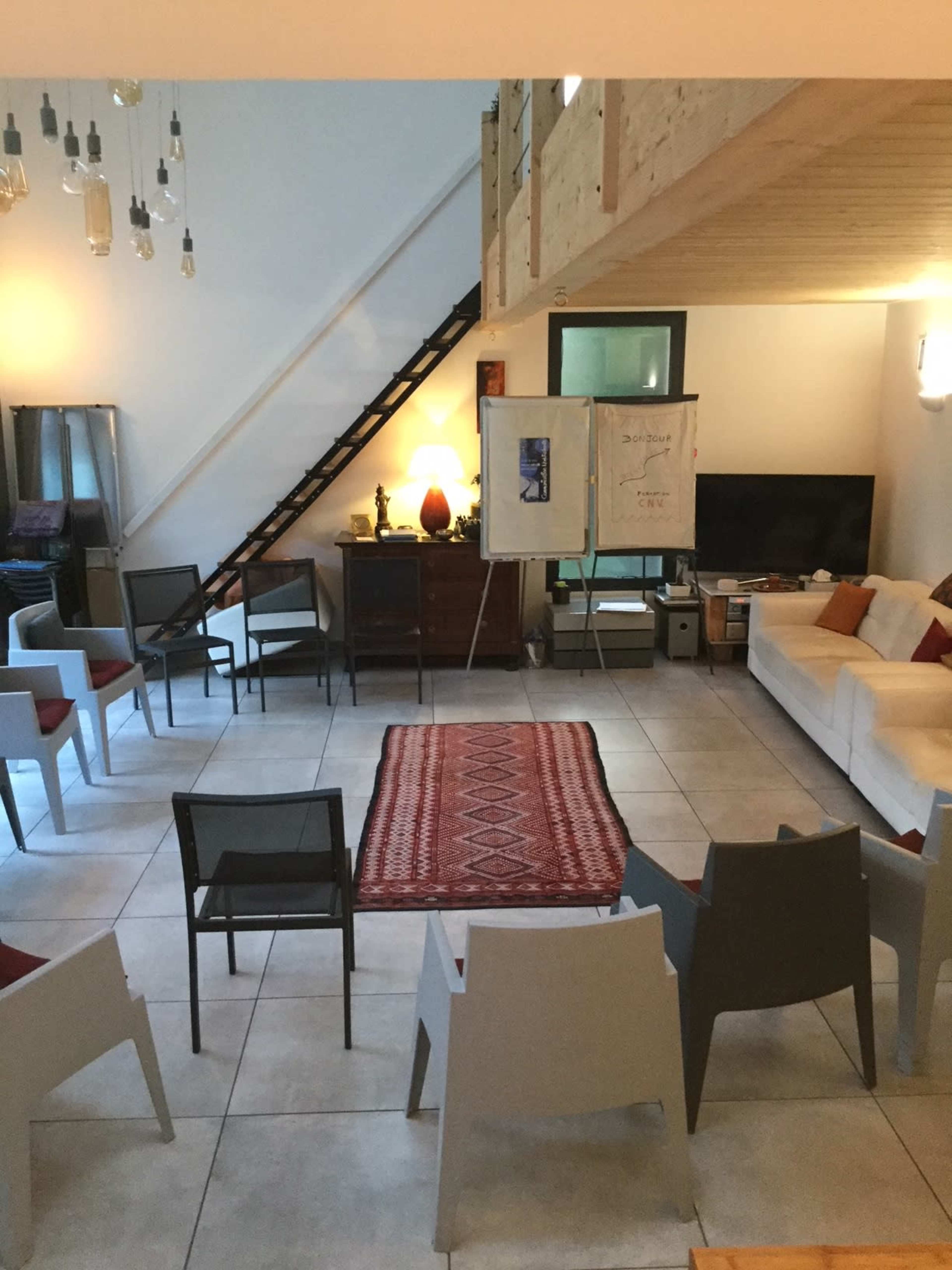 A meeting room with a seating arrangement of several chairs arranged around a central rug, a whiteboard, and a large television, all in a bright, open space with a staircase in the background.