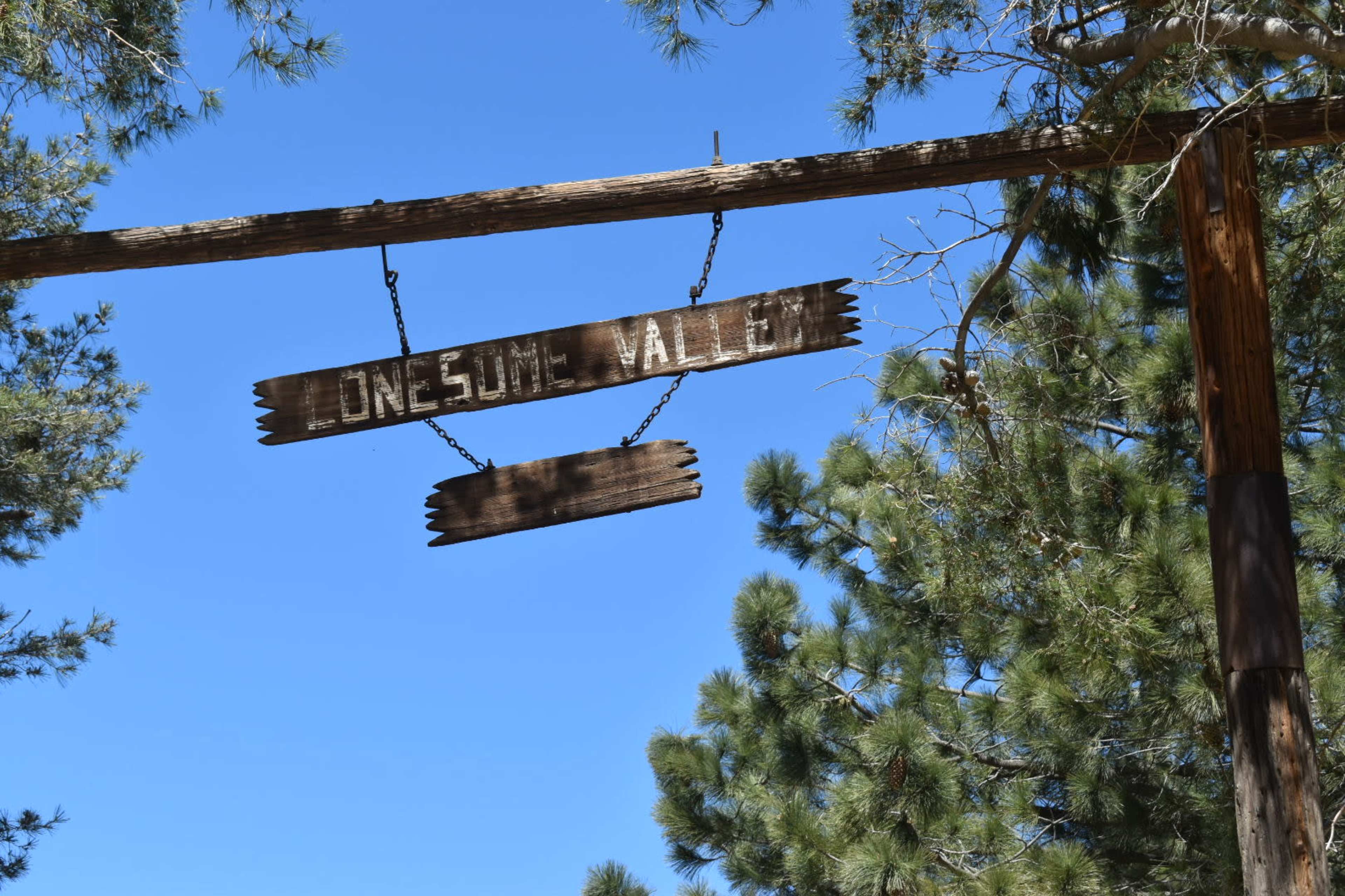 The image shows a weathered wooden sign hanging from a post, reading "Lonesome Valley," amidst tall green trees against a clear blue sky.