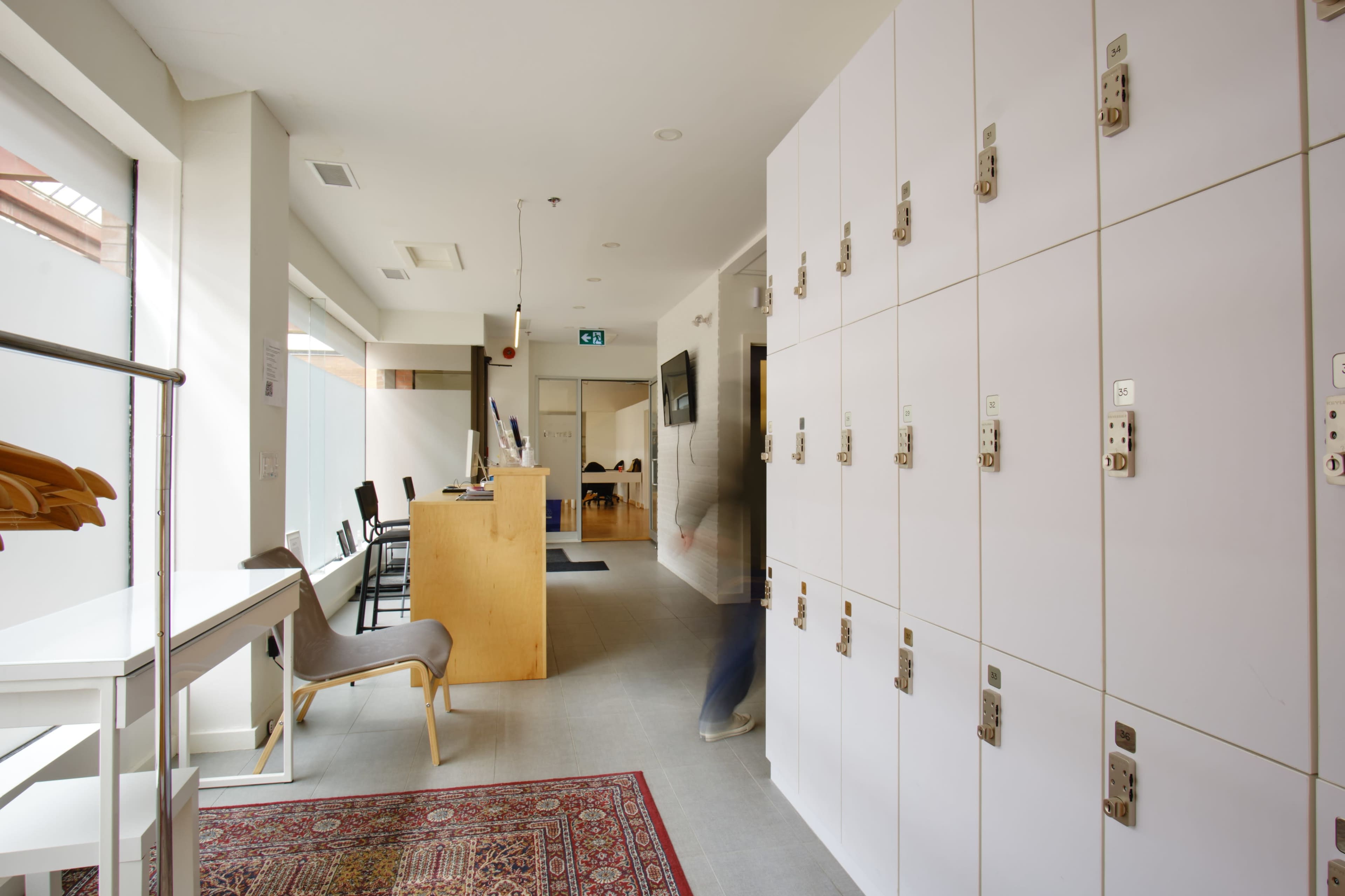 The image shows a modern interior space featuring a reception area with lockers along one wall and a corridor leading to another room.