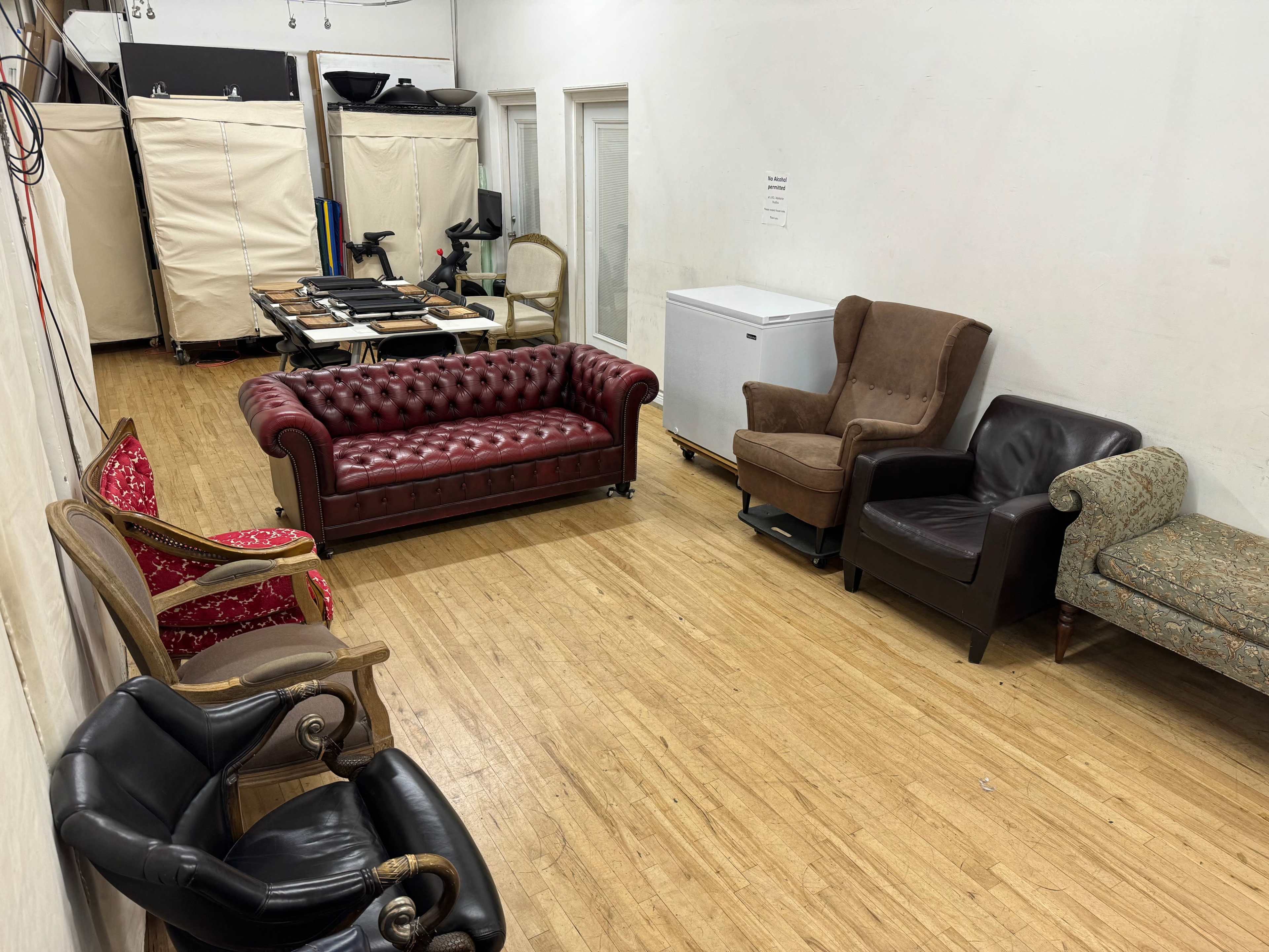 The image shows a room with multiple seating arrangements, including a red tufted sofa, two armchairs, and a patterned chair, against a backdrop of white walls and wooden flooring.
