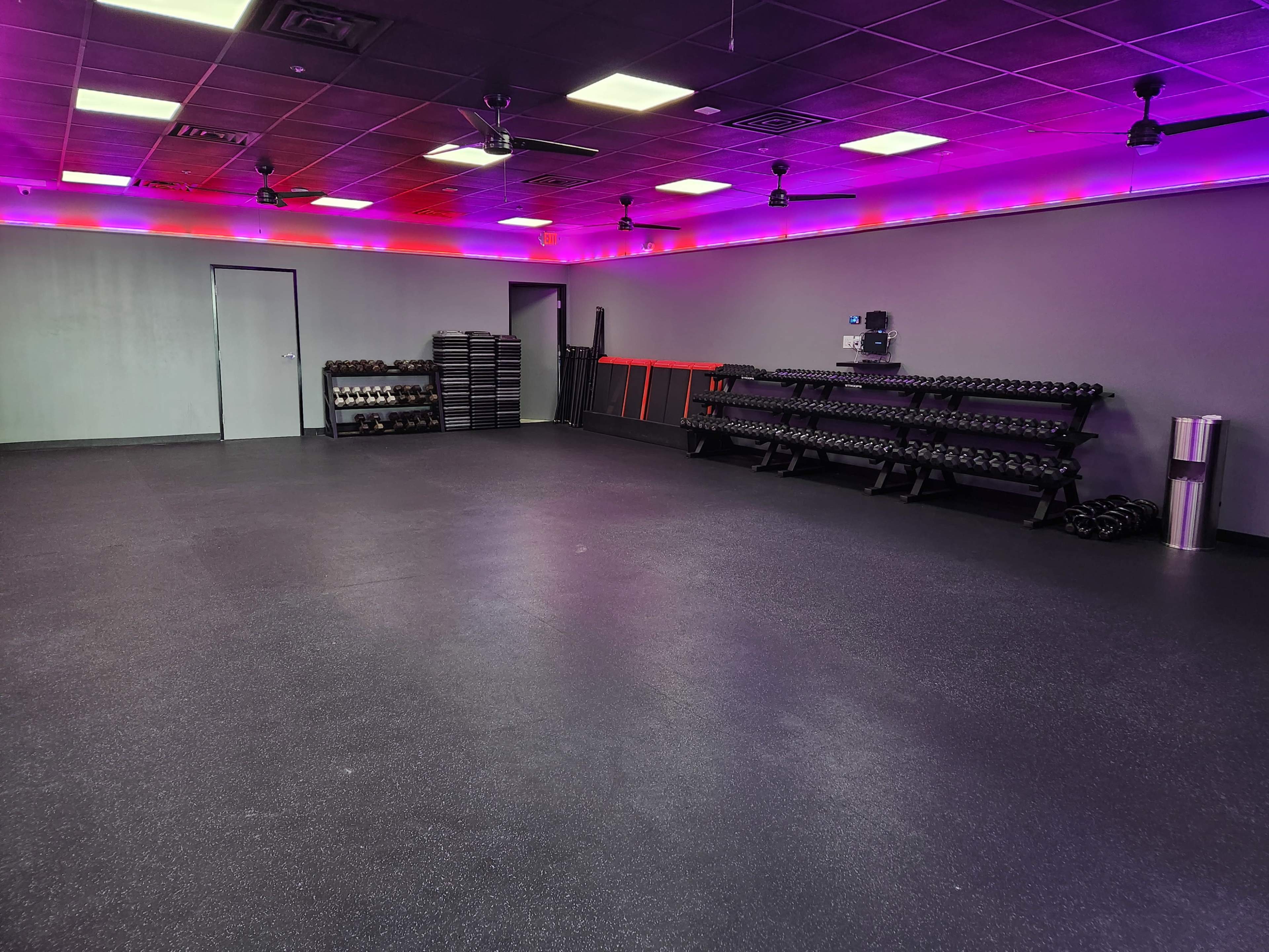 An empty gym space with various weights organized on racks along the walls, illuminated by purple lighting.