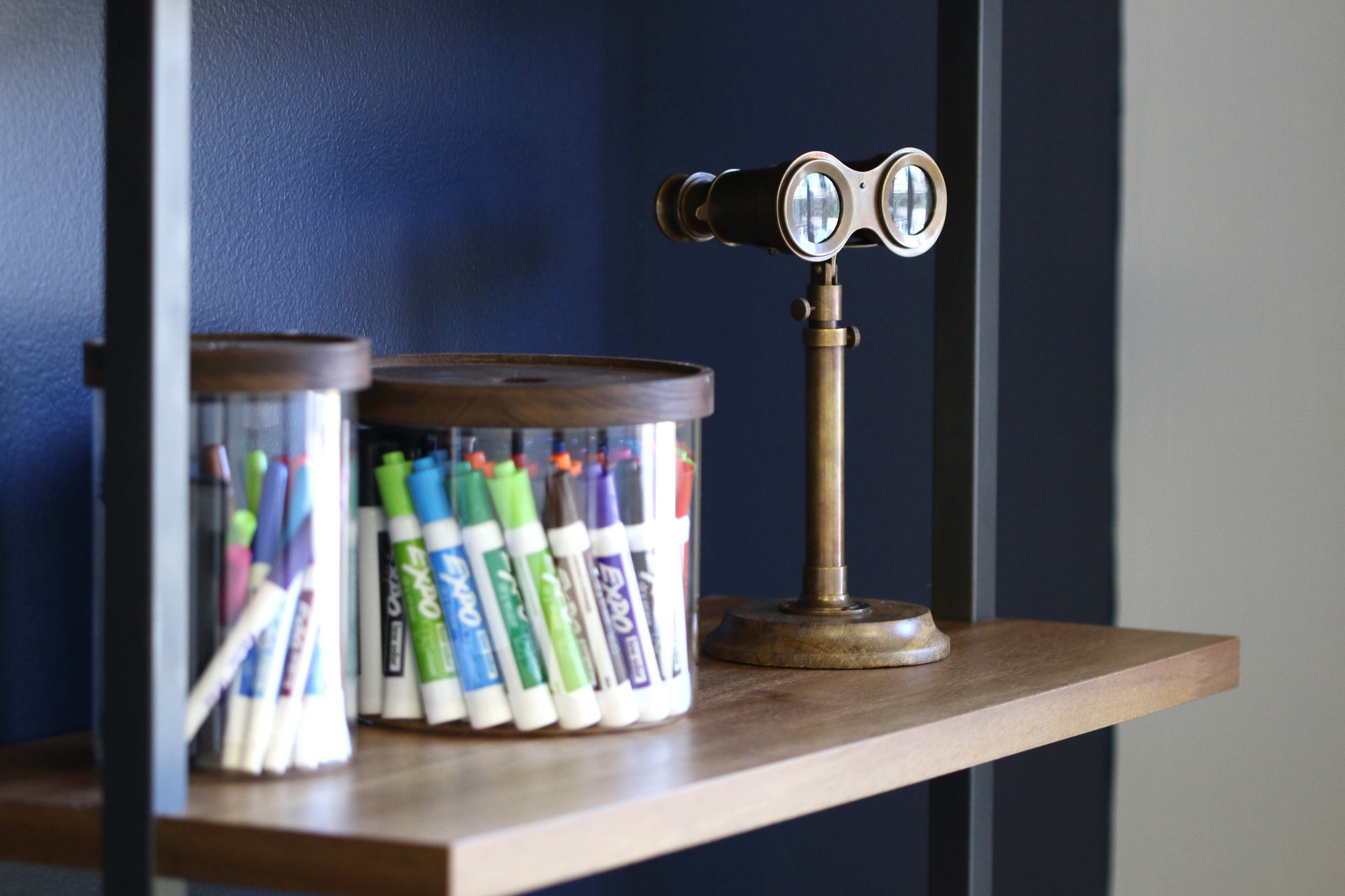 A wooden shelf displays two transparent containers filled with markers and a small vintage binoculars stand.