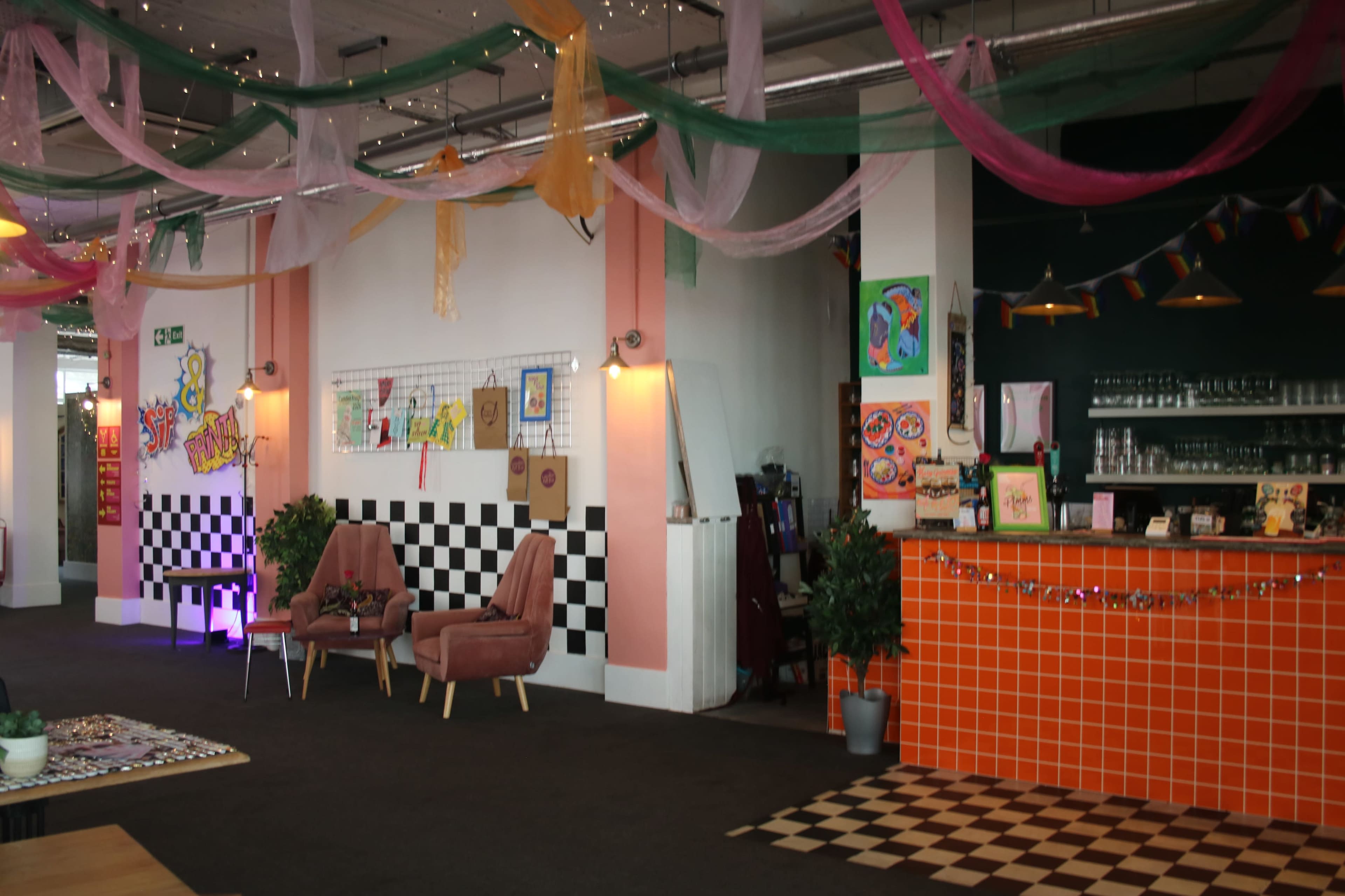 The image shows a vibrant interior of a café or creative space, featuring checkered flooring, colorful artwork on the walls, and cozy seating arrangements.