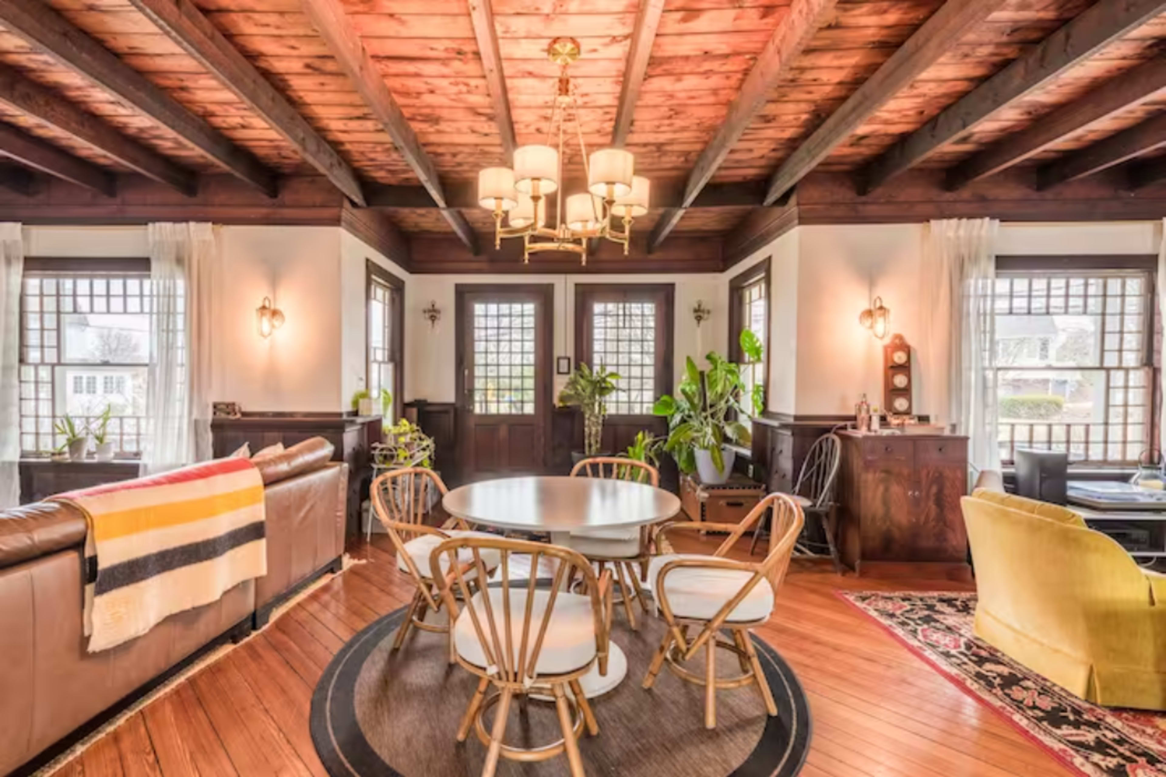 The image shows a dining area with a round table surrounded by light-colored chairs, under a chandelier, in a room featuring wooden beams and large windows.