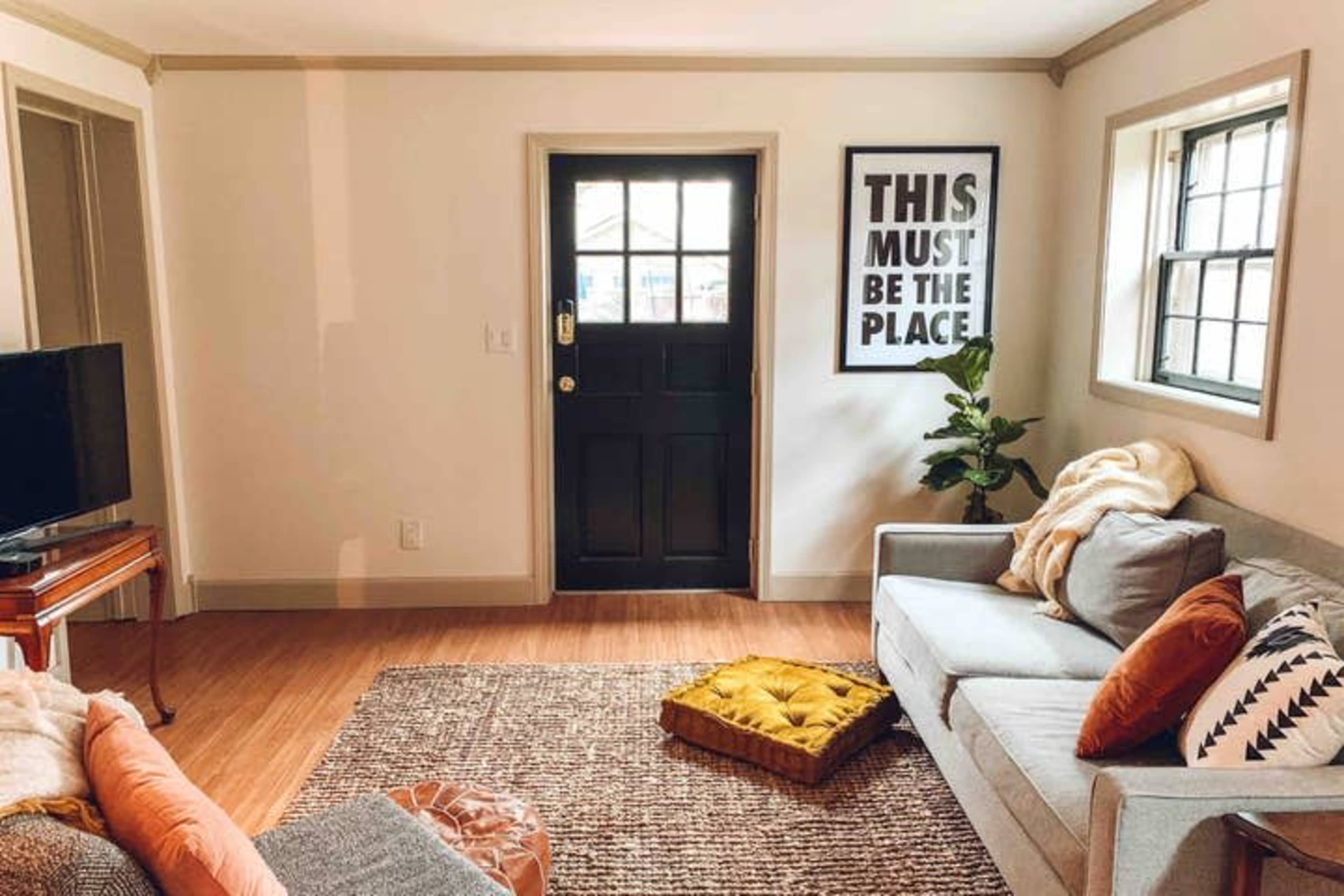 A living room features a gray sofa, an orange throw pillow, a yellow cushion on the floor, a flat-screen TV, and a door with a large framed poster beside it.