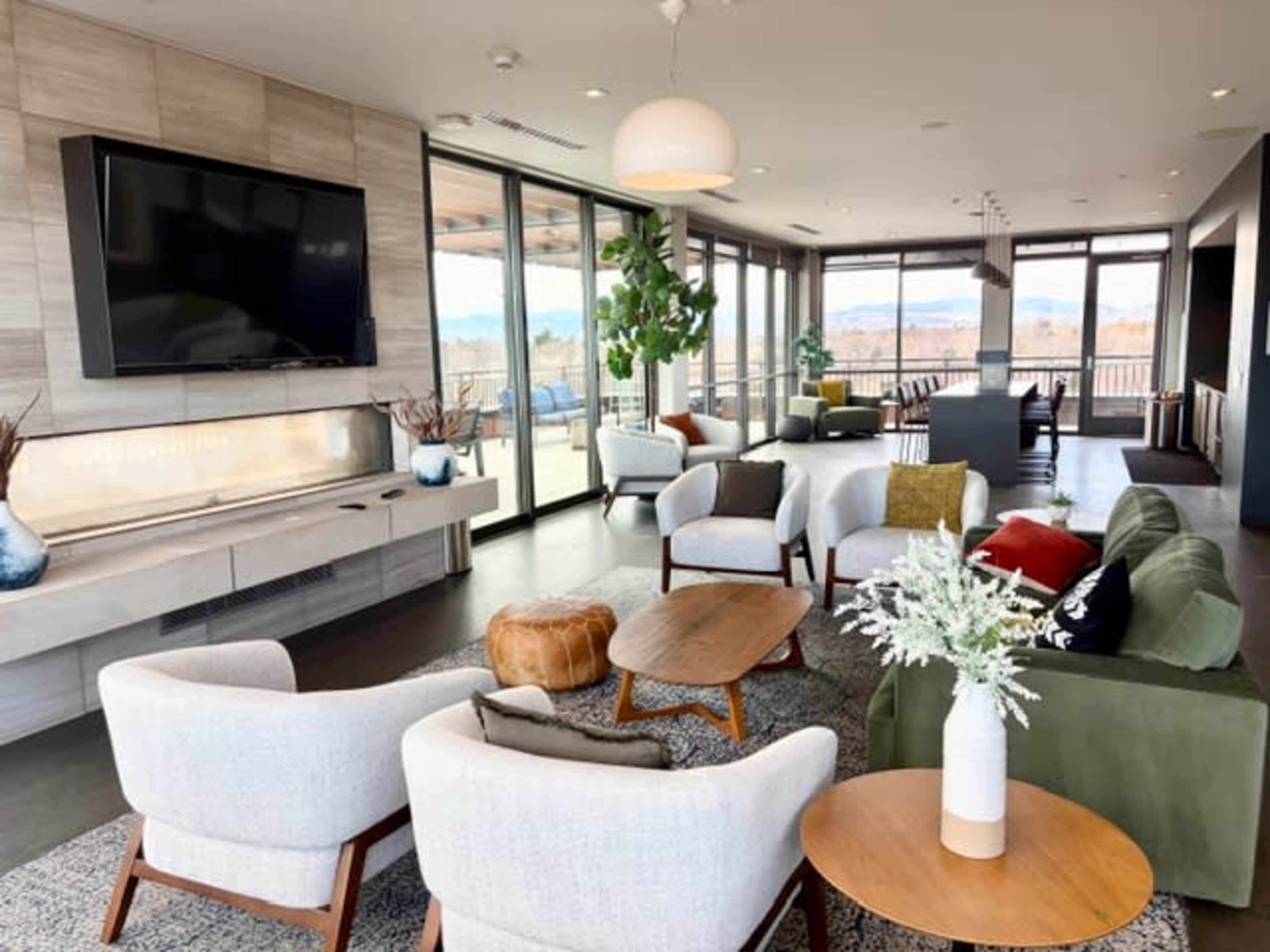 The image shows a modern lounge area with large windows, comfortable seating arrangements, and a television mounted on the wall.