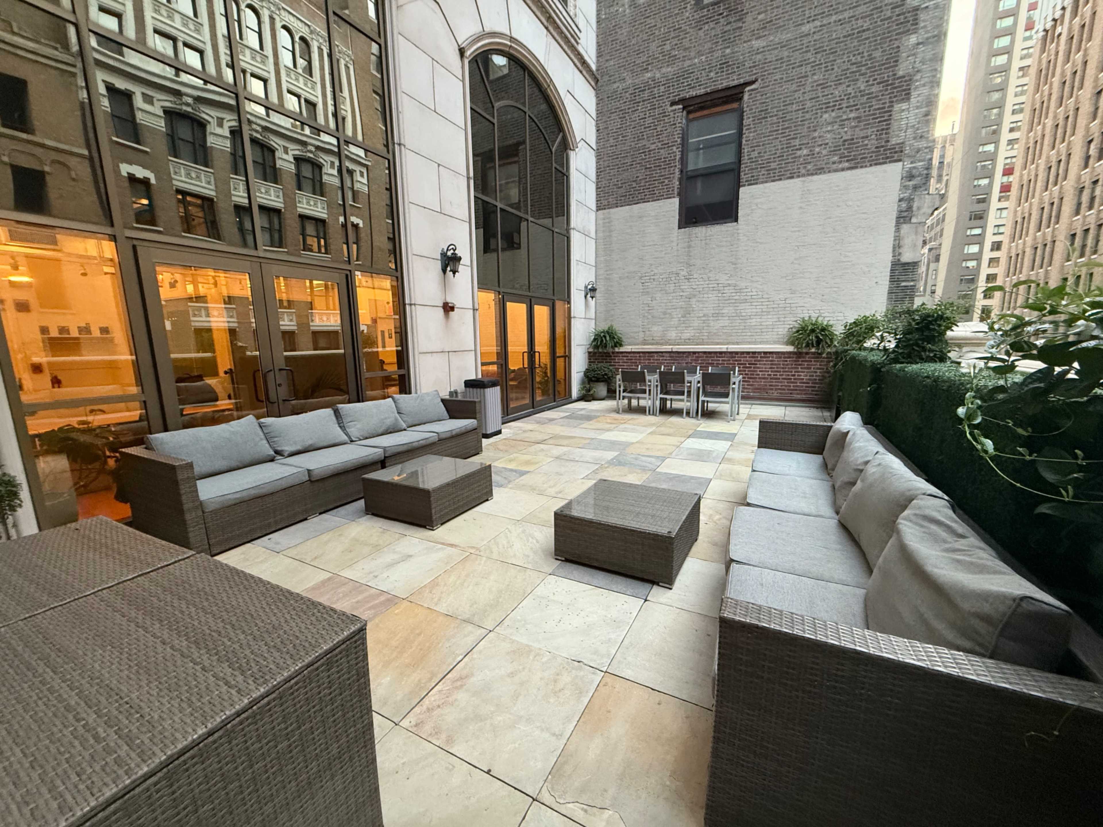 The image shows a quiet outdoor patio with wicker furniture, including sofas and tables, surrounded by tall buildings.